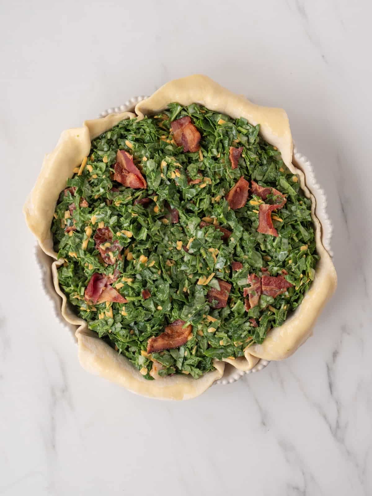 A 9 inch pie dish with rolled out puff pastry, filled with a mixture of eggs, milk, spinach, bacon and cheese, ready to be baked in the oven.