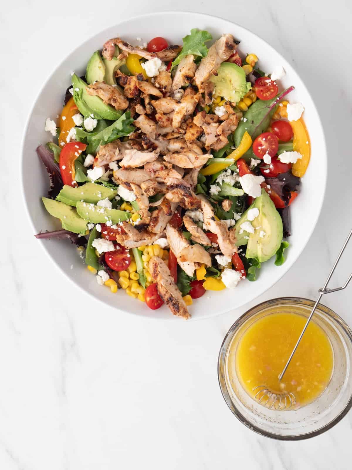 A white low bowl with chicken fajita cobb salad, with a small glass bowl of vinaigrette with a tiny whisk.