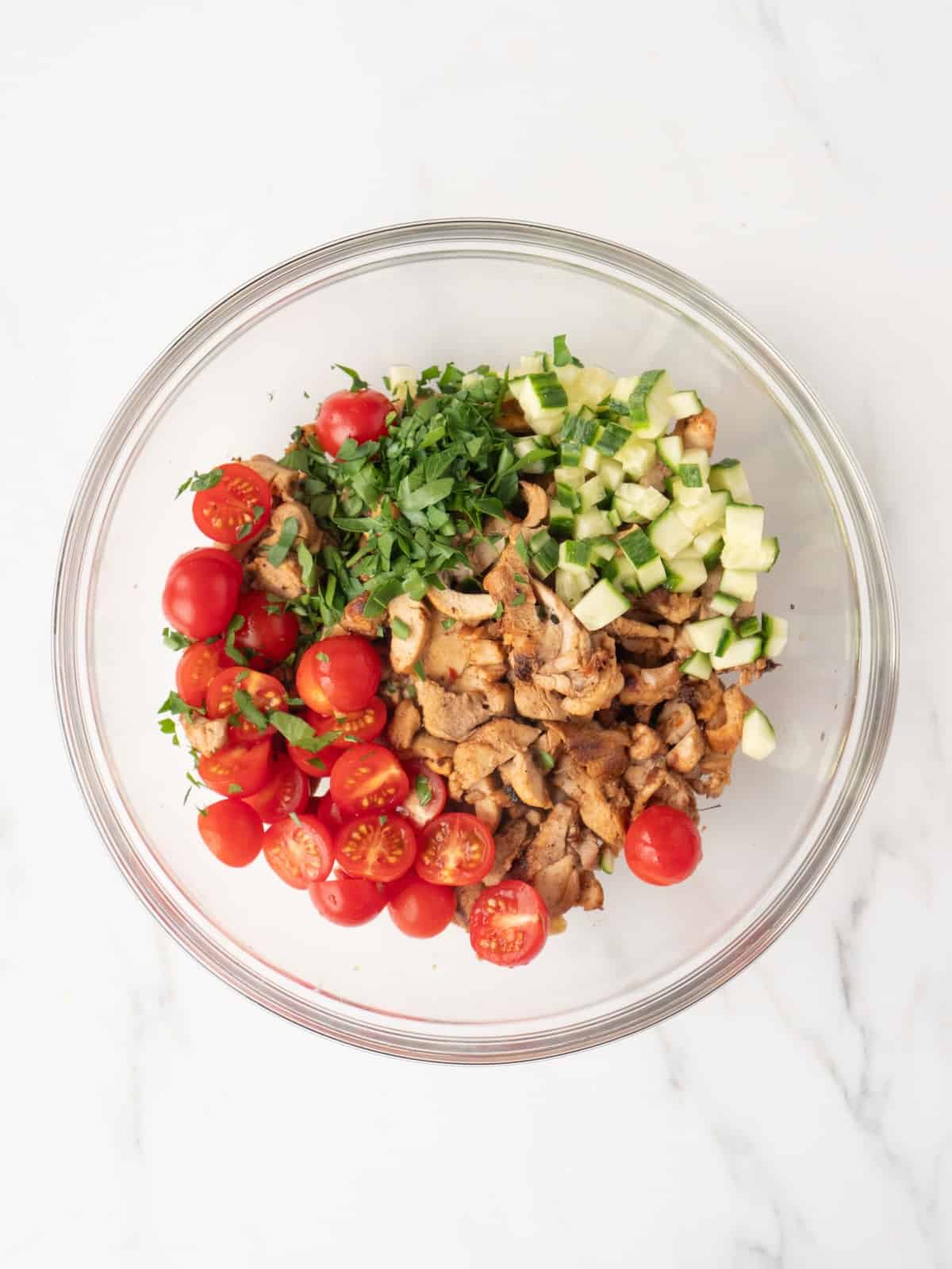 A large glass mixing bowl with grilled sliced chicken, halved cherry tomatoes, chopped cucumbers and parsley.