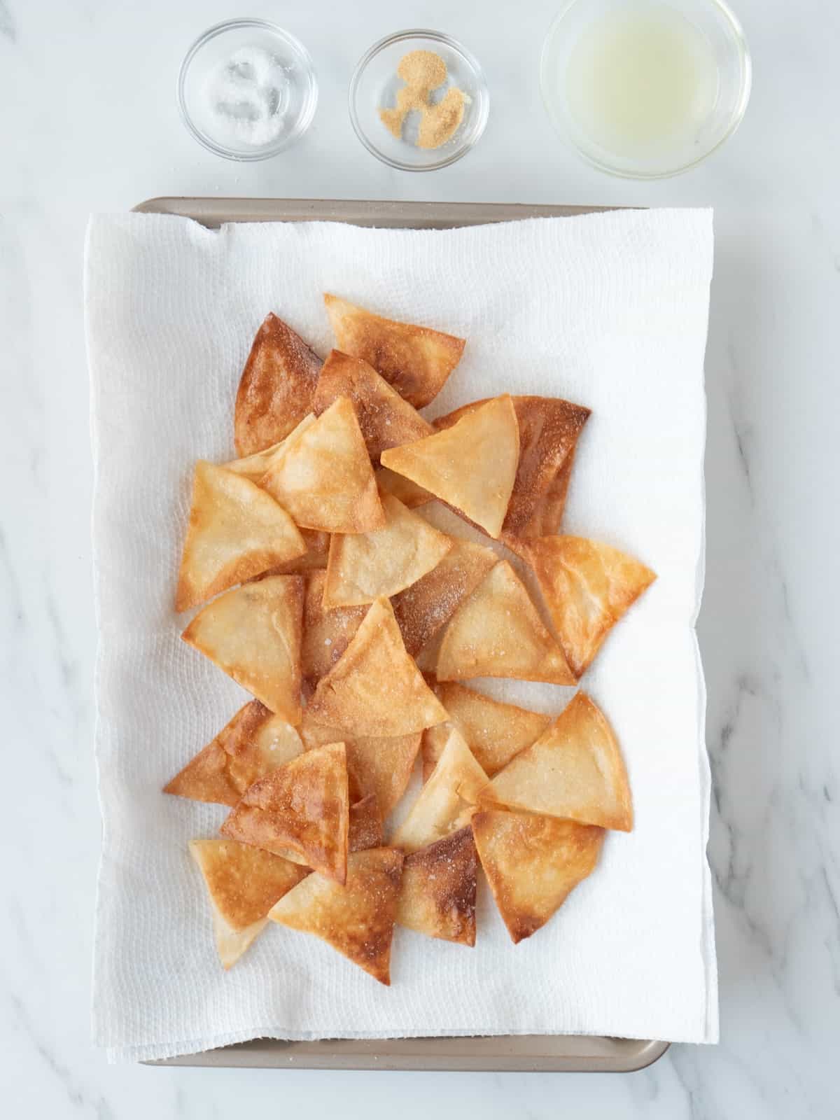 A baking sheet lined with tissues with freshly fried tortilla chips, with three small glass mixing bowls of salt, garlic powder and lime juice on the side.