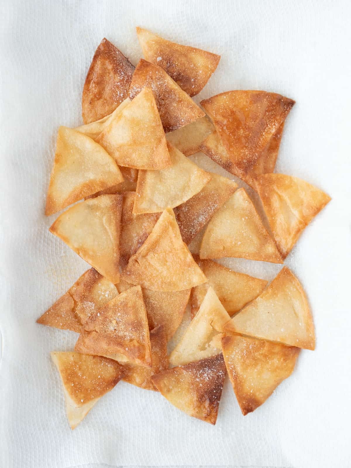 A close up shot of fried tortilla chips on white tissues to soak the excess oil, the chips are sprinkled with salt and garlic powder.