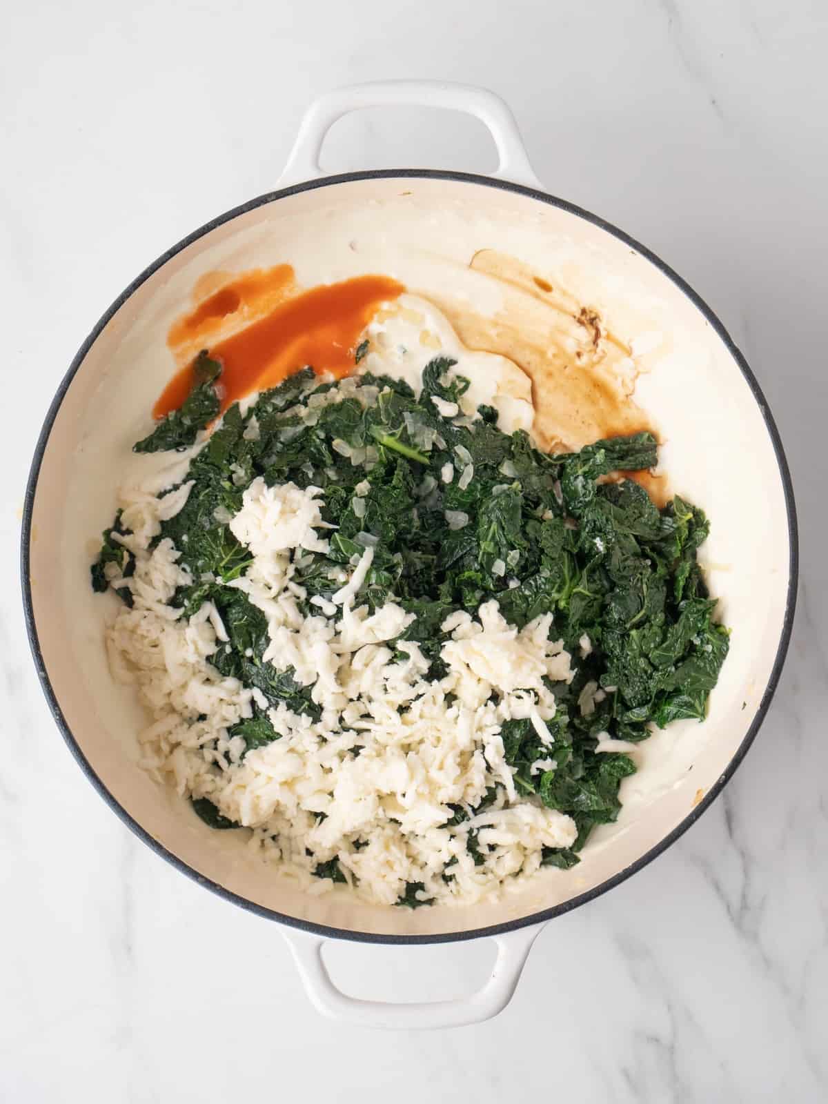 A large white dutch oven with kale, Worcestershire sauce, hot sauce, and the mozzarella added to a milk and cream cheese mixture.