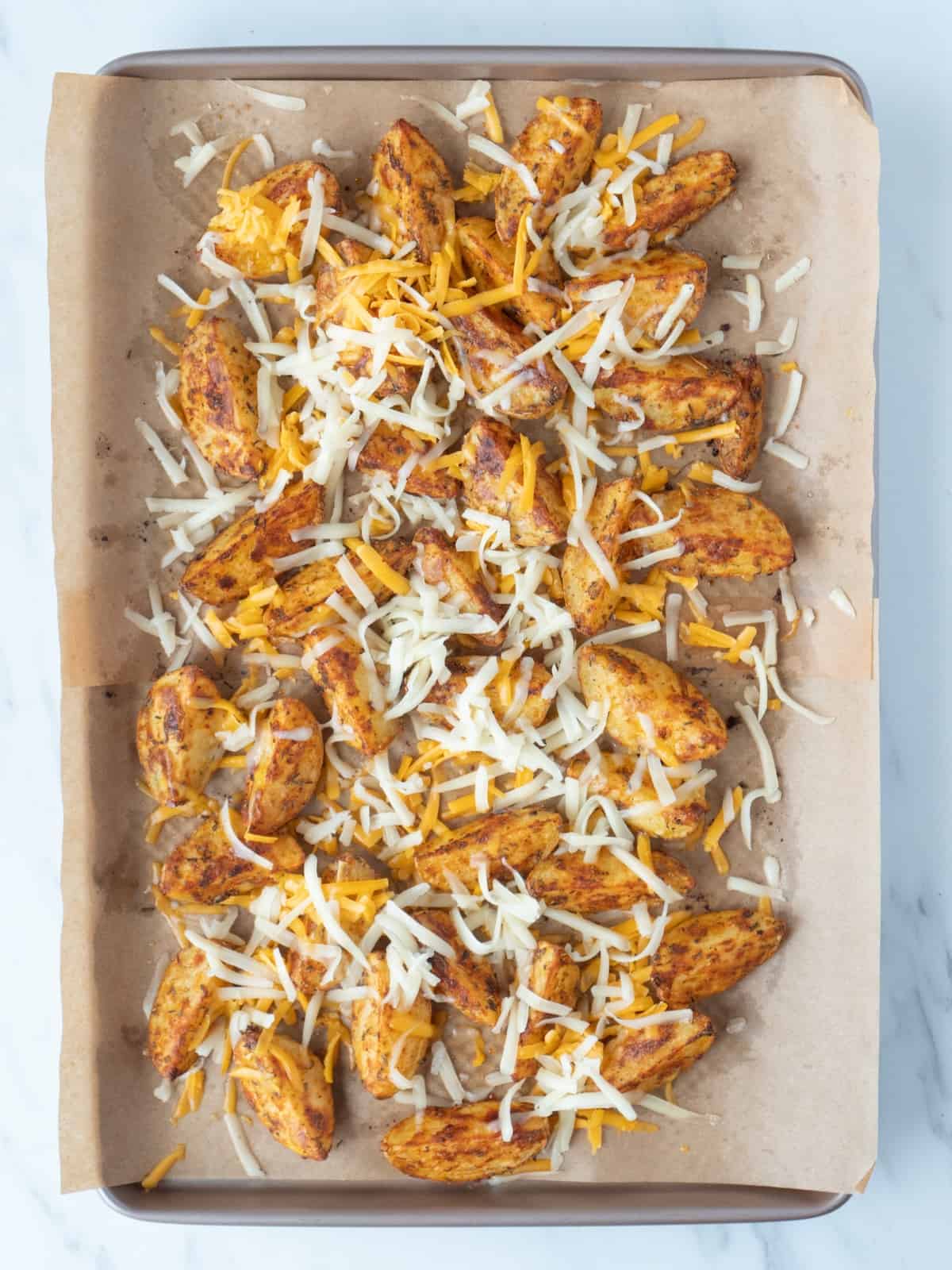 A parchment-lined baking sheet with quartered potatoes, roasted in the oven, topped with shredded cheese.