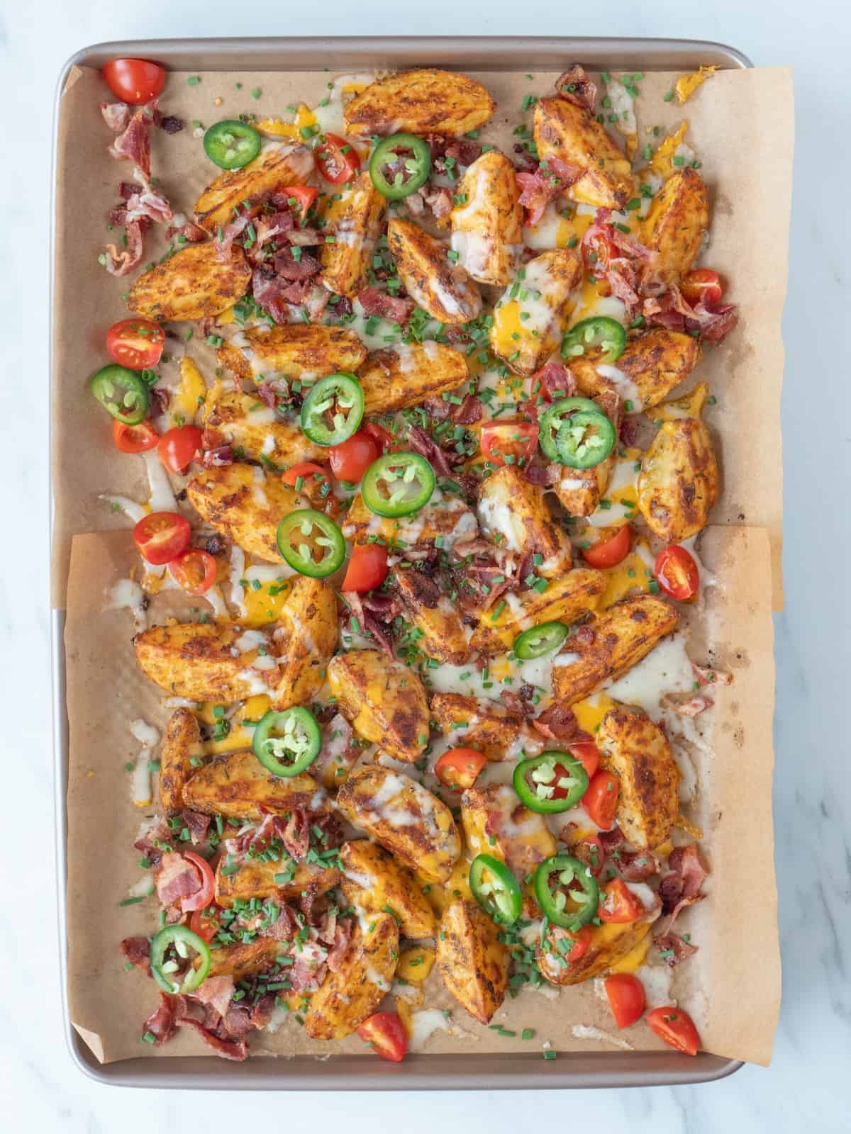 A parchment-lined baking sheet with quartered potatoes, roasted in the oven, covered in a thin layer of melted cheese, topped with crumbled bacon, chopped tomatoes, and sliced jalapeños.