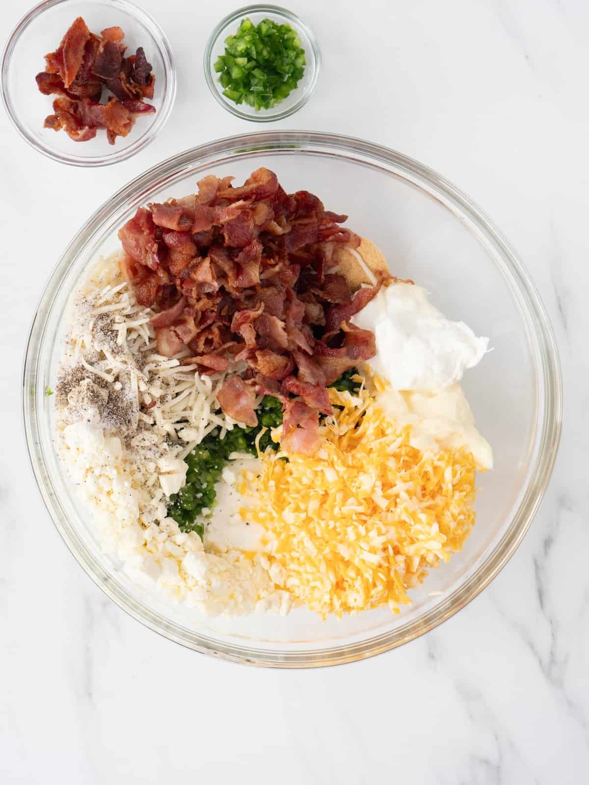 A large glass mixing bowl with all the ingredients to make jalapeño popper dip, with two small glass mixing bowls with bacon bits and chopped scallions.