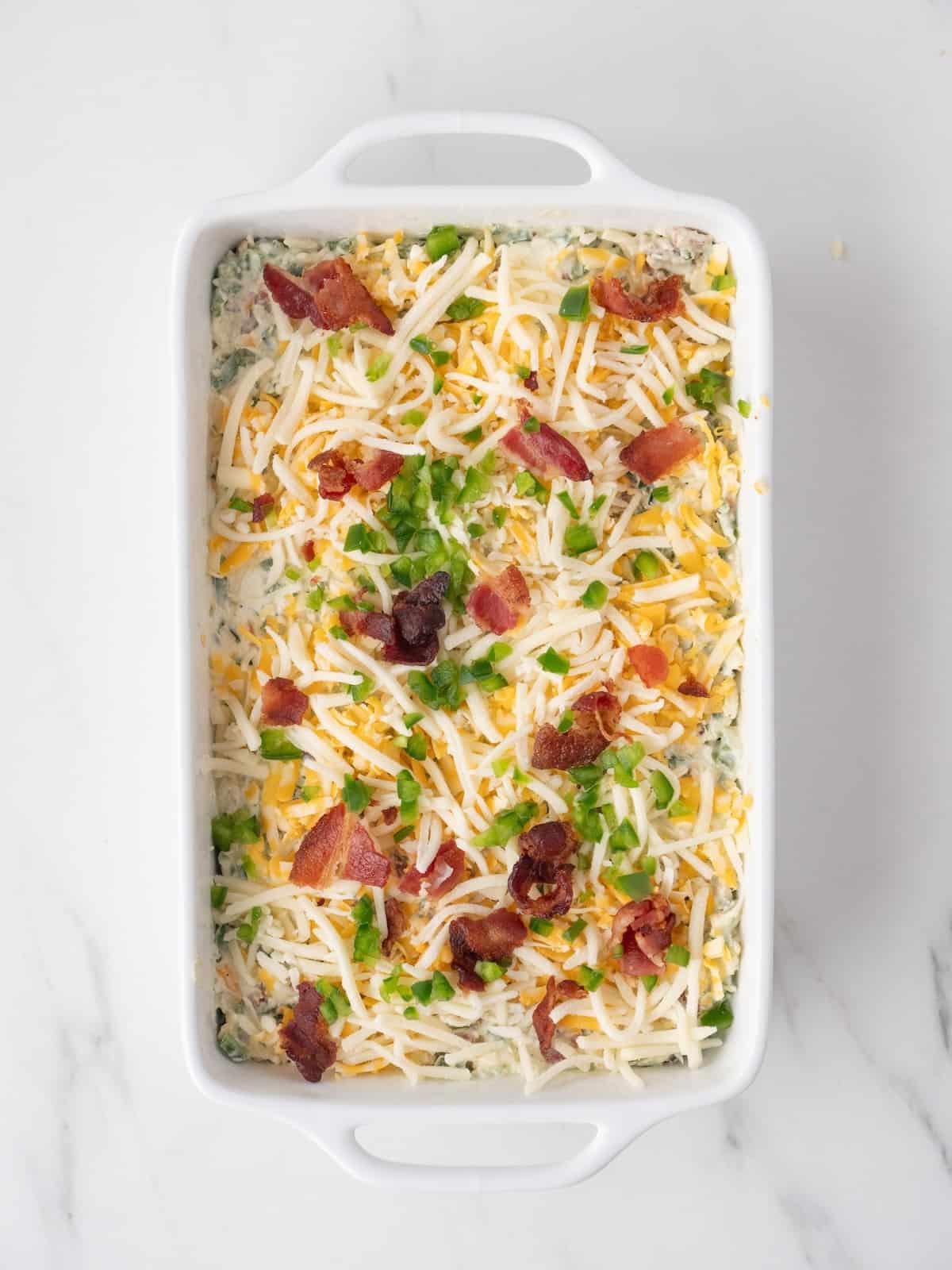 A rectangular white dish with the jalapeño popper dip mix sprinkled with shredded cheese, crumbled bacon and jalapeños on top, ready to go into the oven.