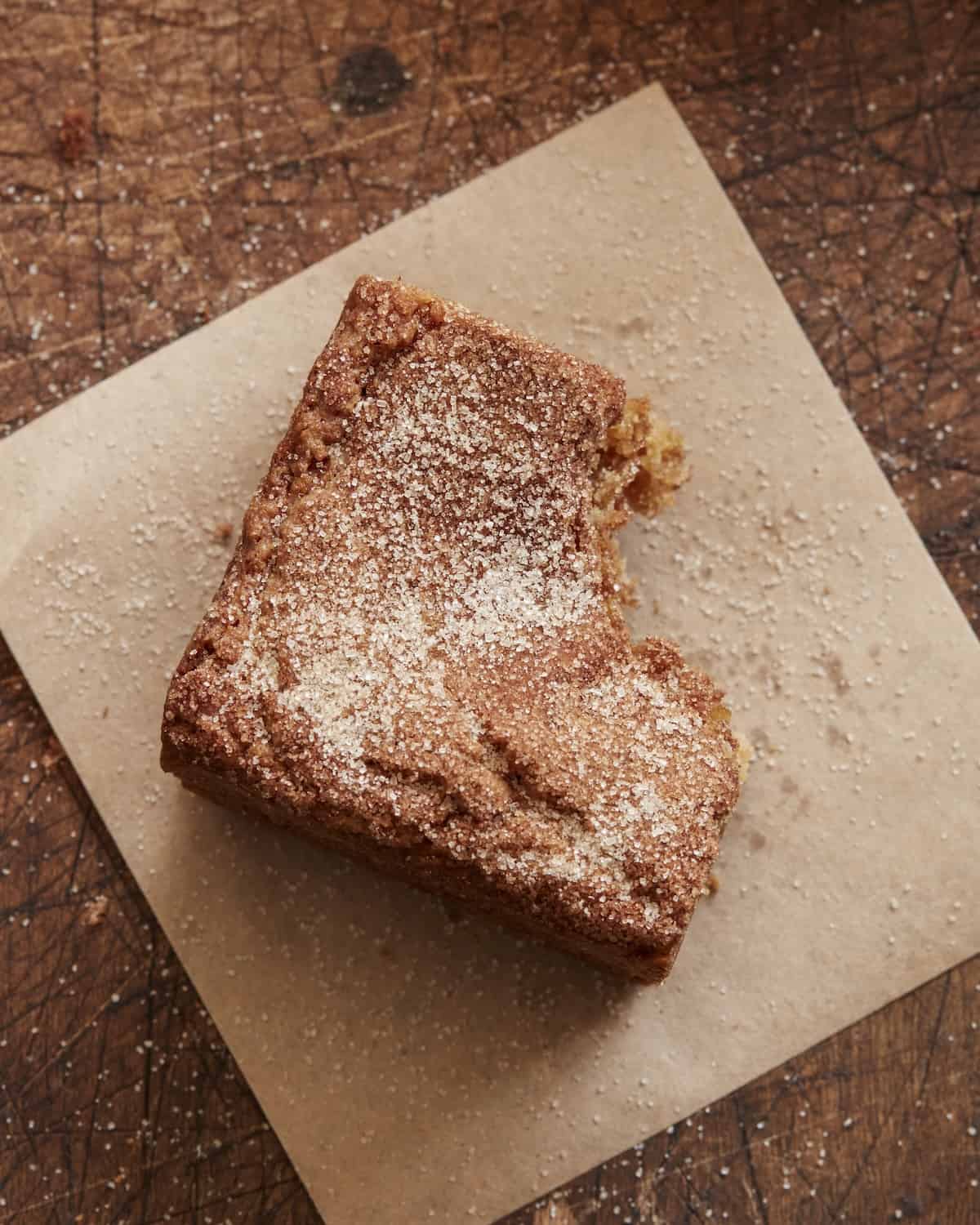 A blondie square placed on a piece of parchment paper, sprinkled with cinnamon sugar and a bite eaten off from the corner, placed on a wooden board.
