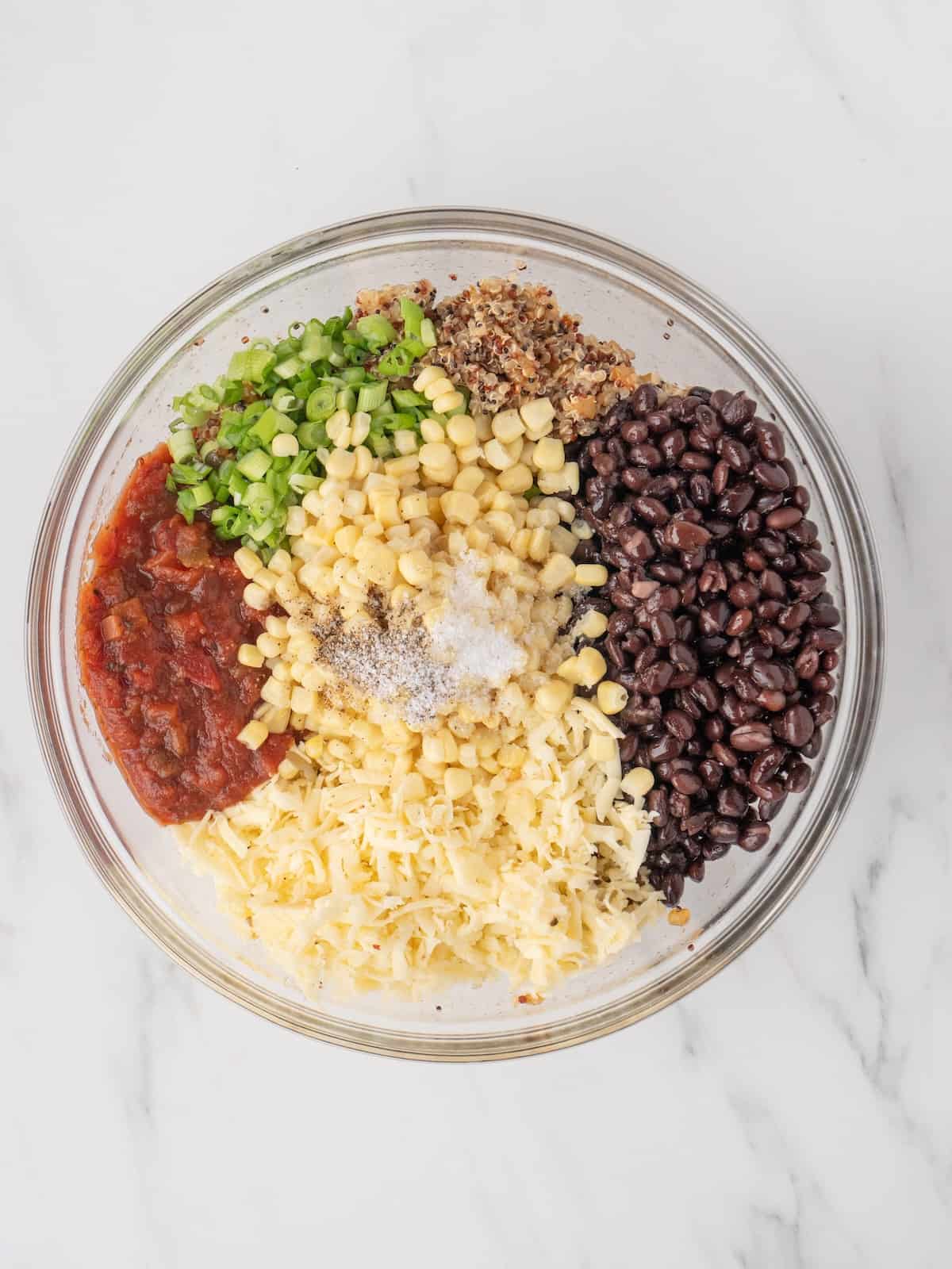 A large glass mixing bowl with corn, black beans, quinoa, shredded cheese, green onions and salsa, topped with salt and black pepper.
