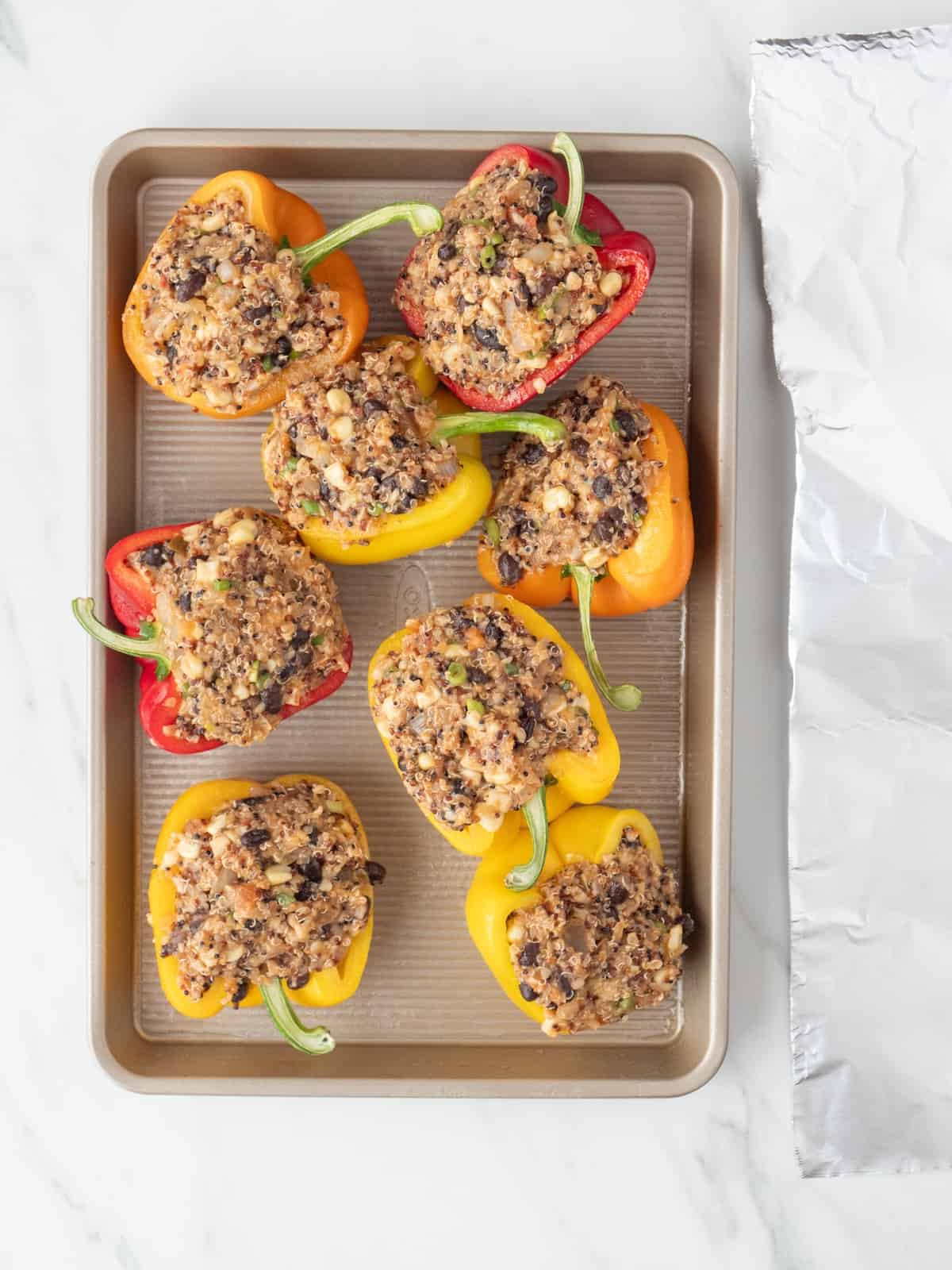 A rectangular baking sheet with halved bell peppers filled with the quinoa, black bean, corn mix, with a piece of aluminium foil on the side.