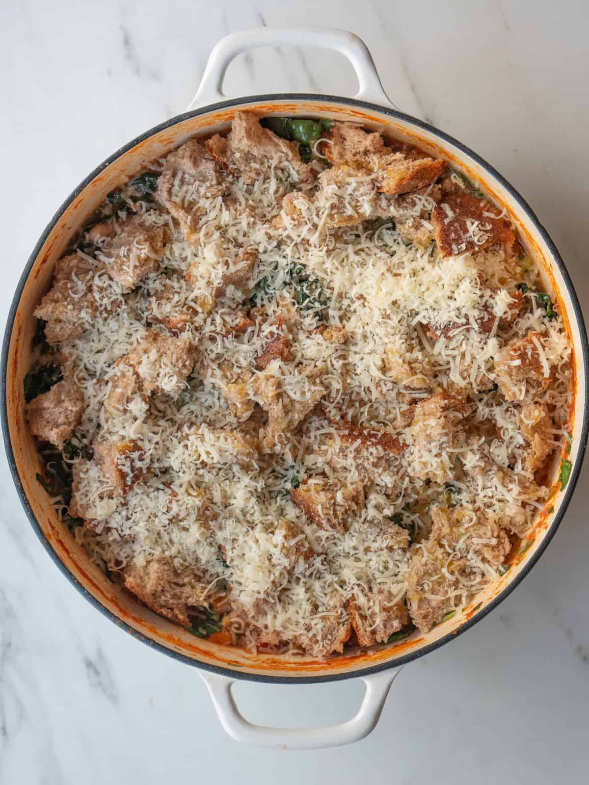 A dutch oven with ribollita soup being made, and bread pieces laid on top in an even layer, along with a drizzle of oil and a heavy sprinkle of grated parmesan cheese.