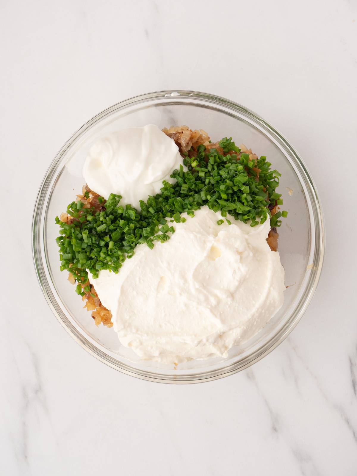 A large glass mixing bowl of roasted onions and shallots, topped with sour cream, yogurt and finely chopped chives. 