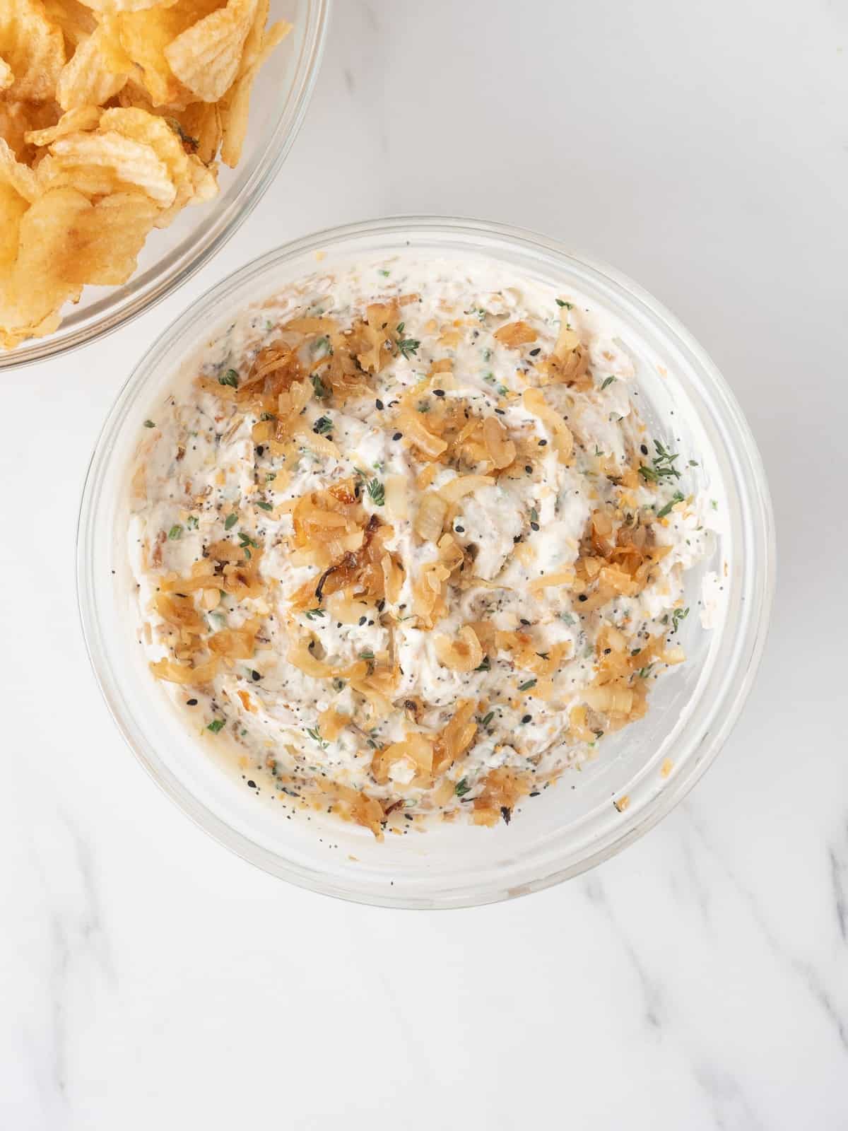 A large glass mixing bowl with roasted onion dip garnished with roasted onion and thyme, and a large bowl of potato chips on the side.