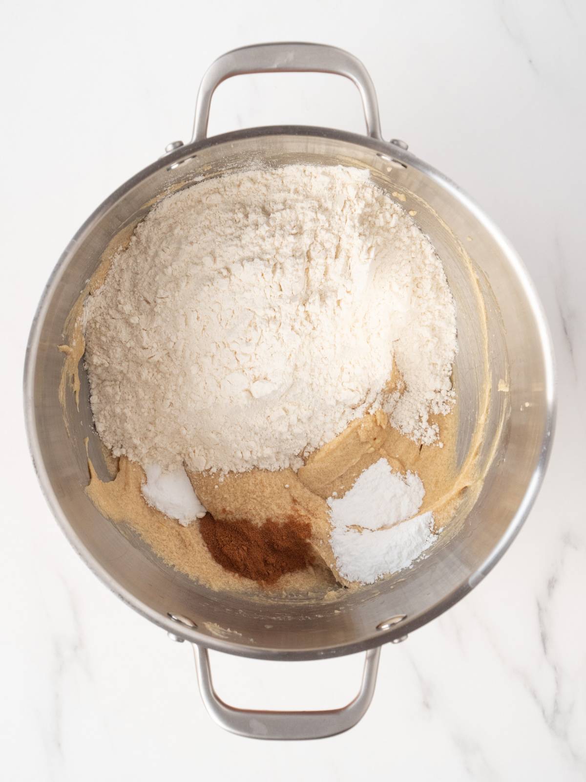 A stand mixer bowl with wet ingredients mixed into a batter, topped with flour, baking powder, baking soda and cinnamon.