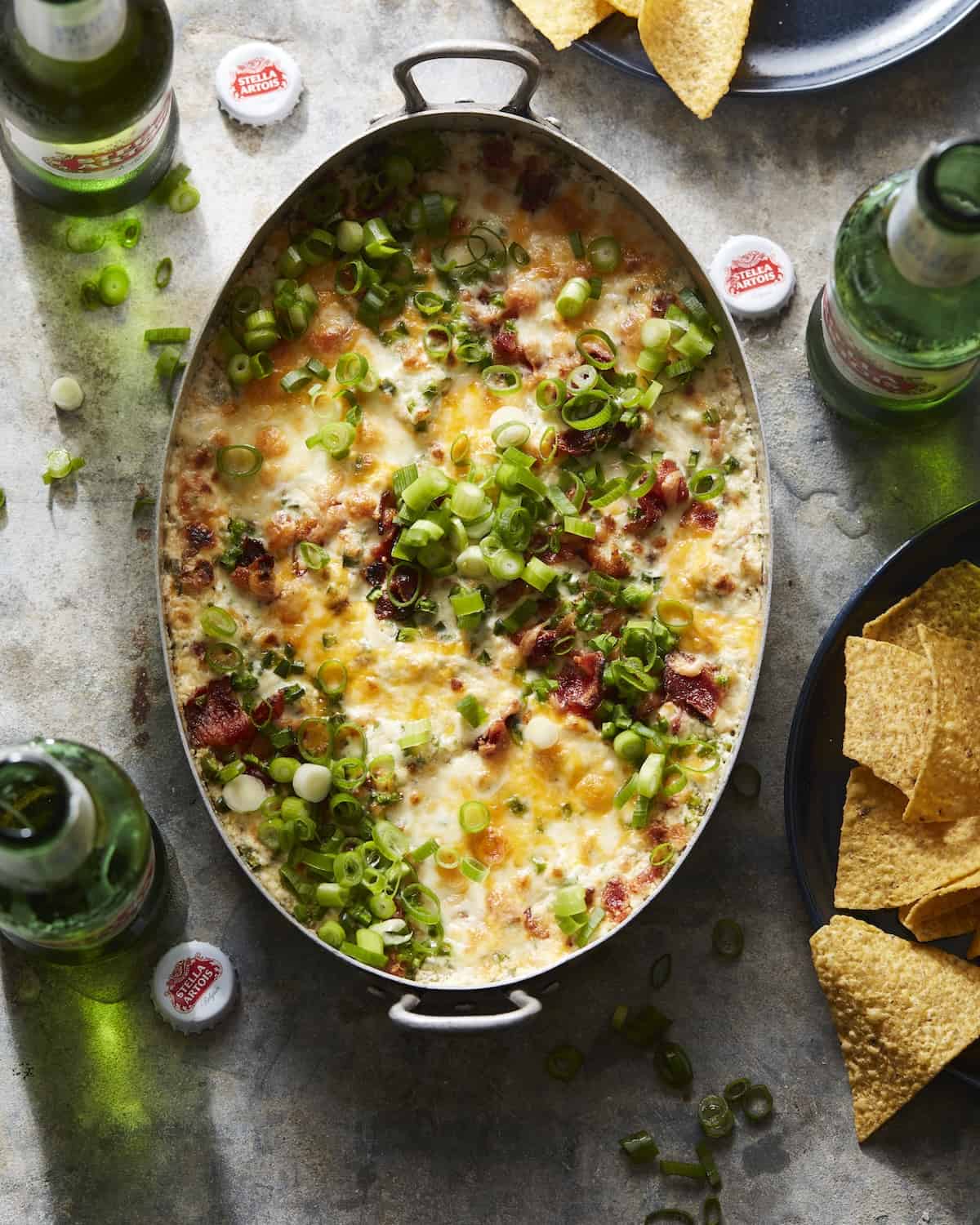 An overheat shot of jalapeño popper dip in a rectangular dish, with Stella Artois beer bottles and plates with tortilla chips on the side.
