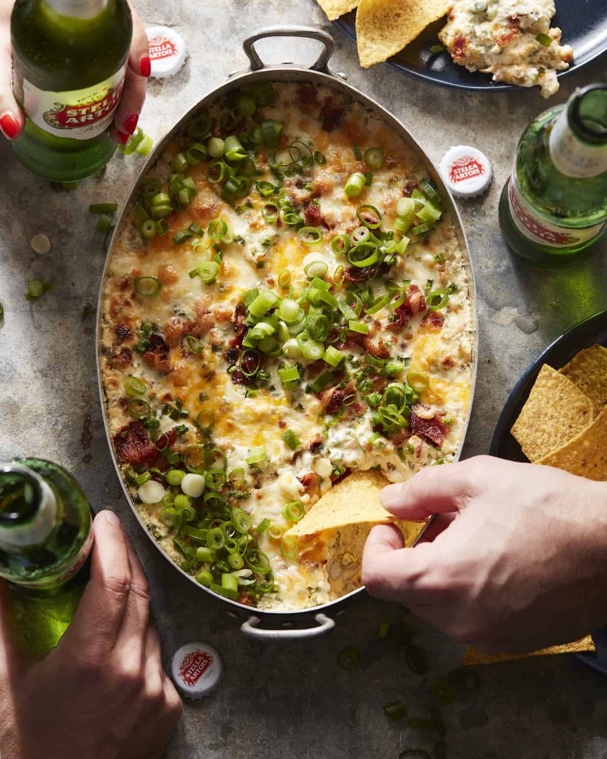 An overheat shot of jalapeño popper dip in a rectangular dish, with someone's hand dipping a tortilla chip into it, and a couple other people's hands holding Stella Artois beer bottles placed on the side.
