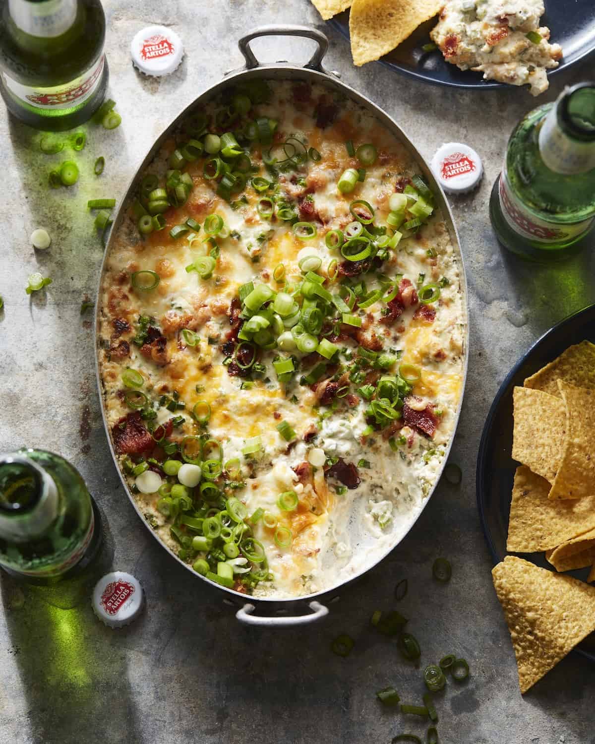 An overheat shot of jalapeño popper dip in a rectangular dish, with Stella Artois beer bottles and plates with tortilla chips on the side.