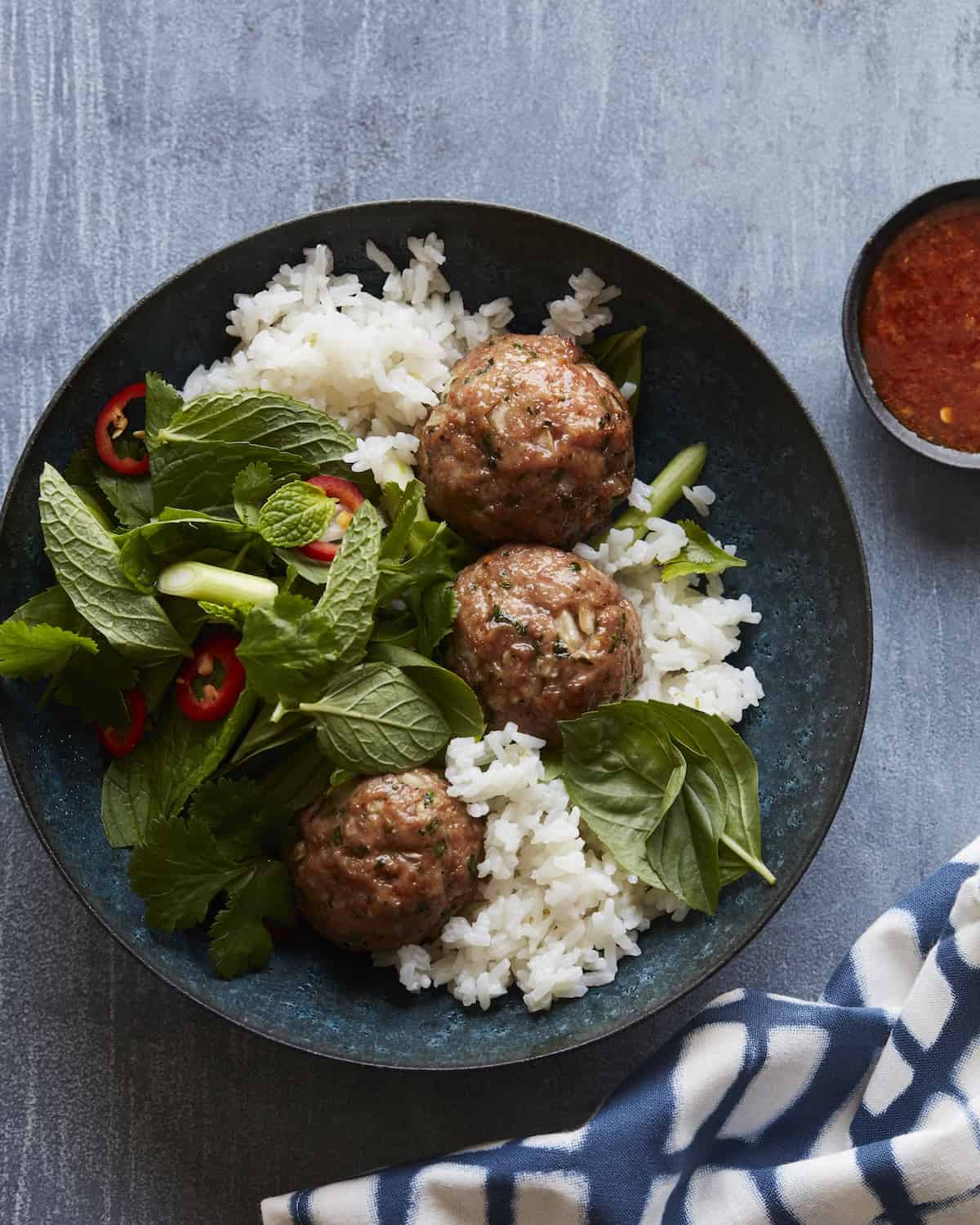 A blue ceramic plate with asian pork meatballs, rice, greens, sliced fresno chilis, a small bowl of sauce, containing soy sauce and sambal, and a kitchen towel on the side.