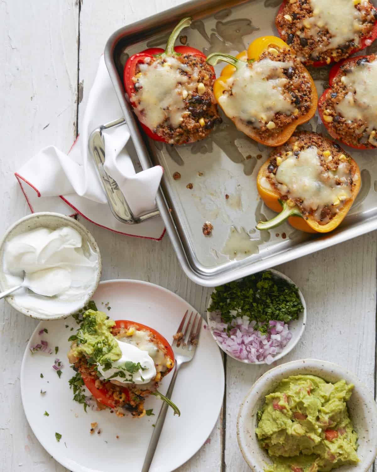 A rectangular deep baking pan with quinoa stuffed peppers, with molten cheese on the top, and three smal bowls of sour cream, guacamole and one with chopped onions and cilantro, on the side, along with a plate and fork with one stuffed pepper served.