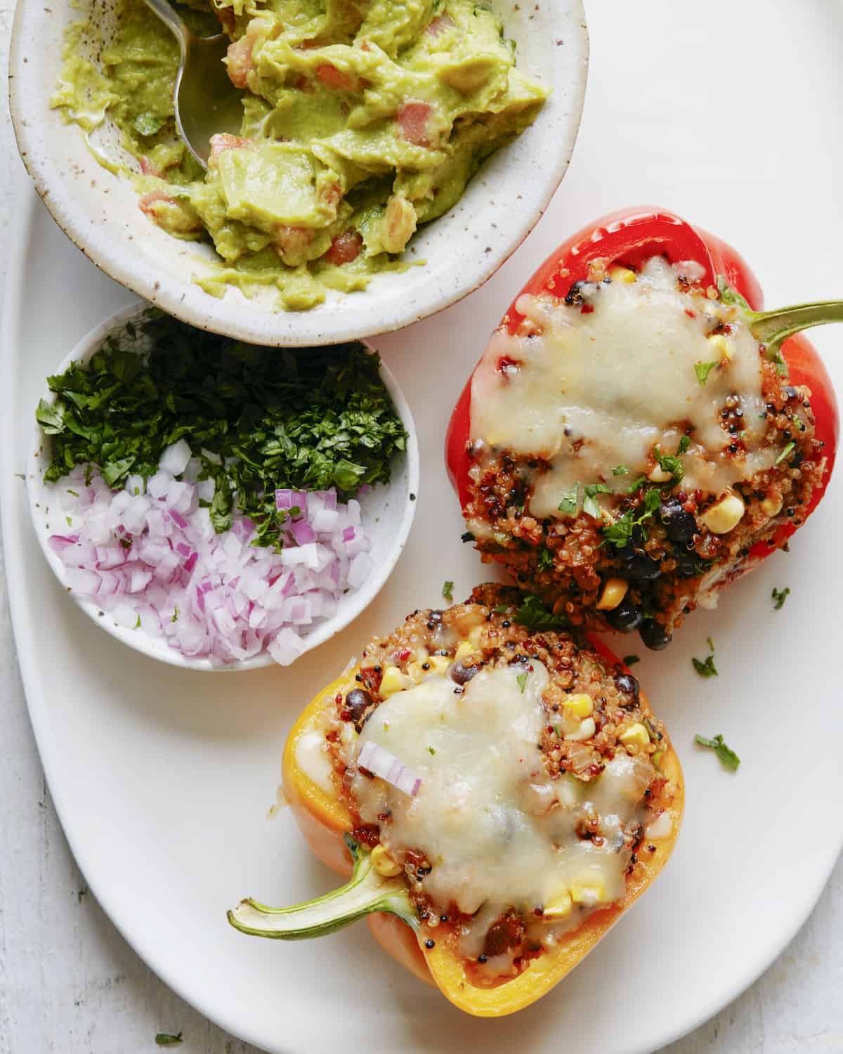 A white plate with quinoa stuffed bell peppers, with a bowl of guacamole and a small bowl of finely chopped onions and cilantro.