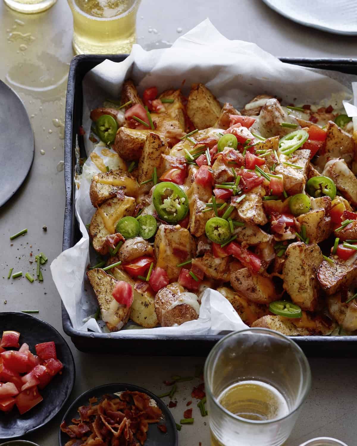 A rectangular baking sheet with jalapeño bacon loaded potatoes, with glasses of beer, and little dishes with chopped tomatoes and crumbled bacon on the side.