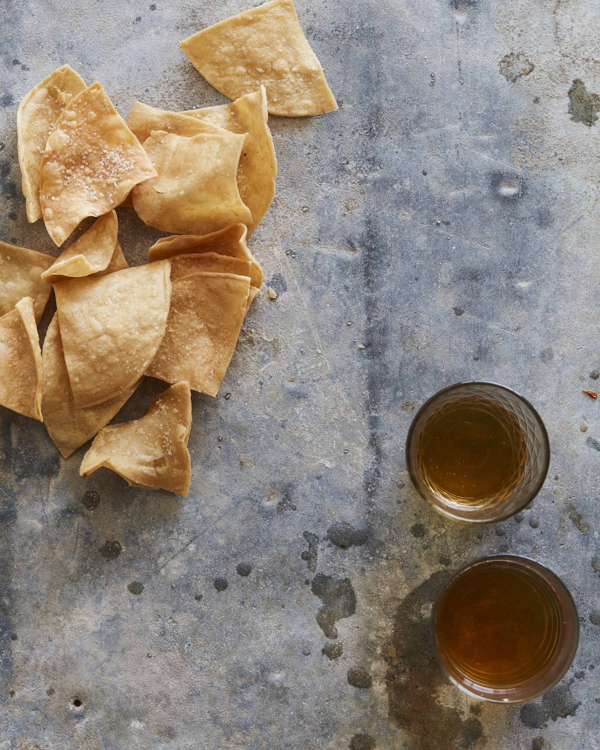 A grey counter with some tortilla chips sprinkled with salt, and two glasses with a liquid in the bottom right.