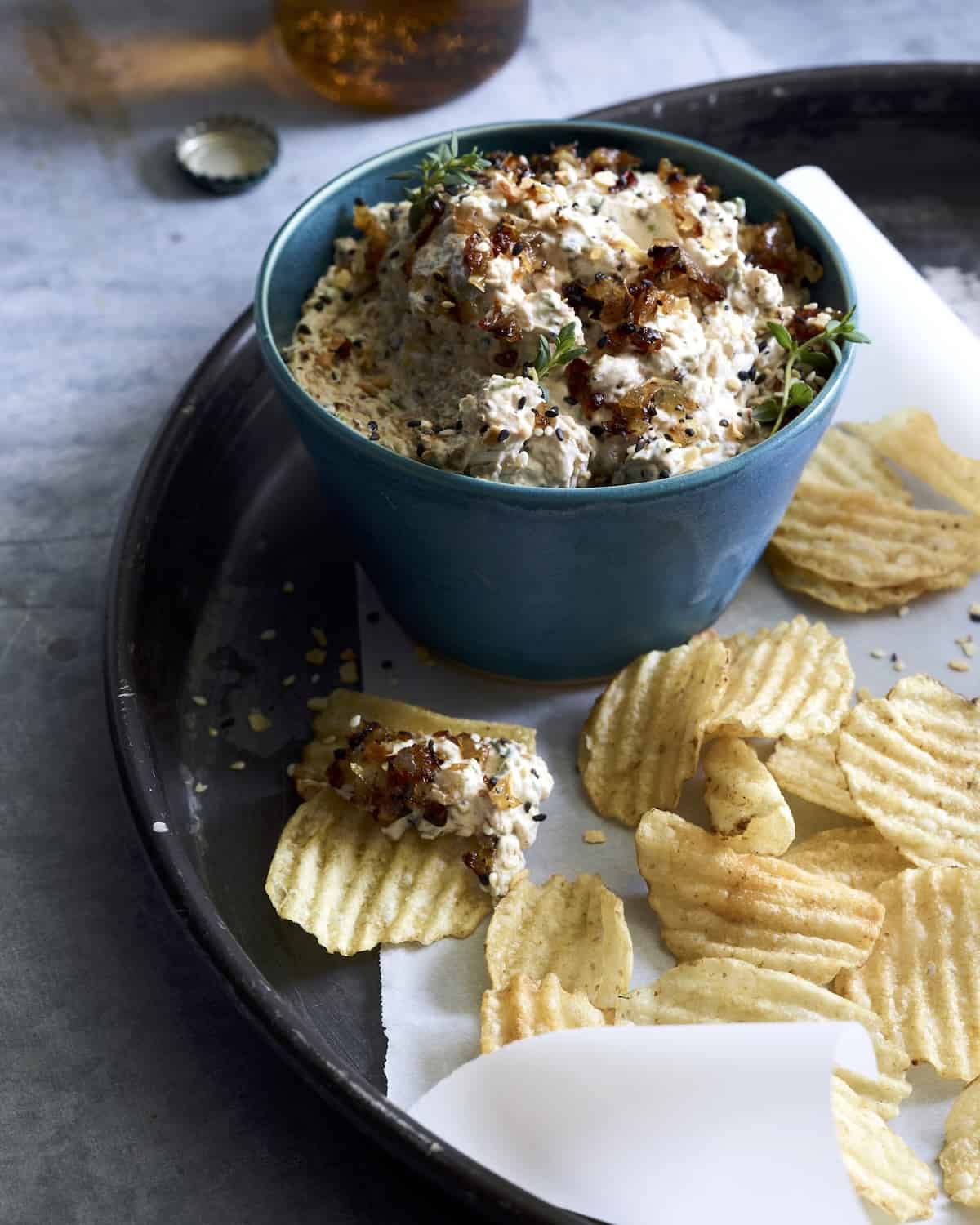 A parchment lined round platter with wavy potato chips, along with a blue bowl of roasted onion dip garnished with thyme. 
