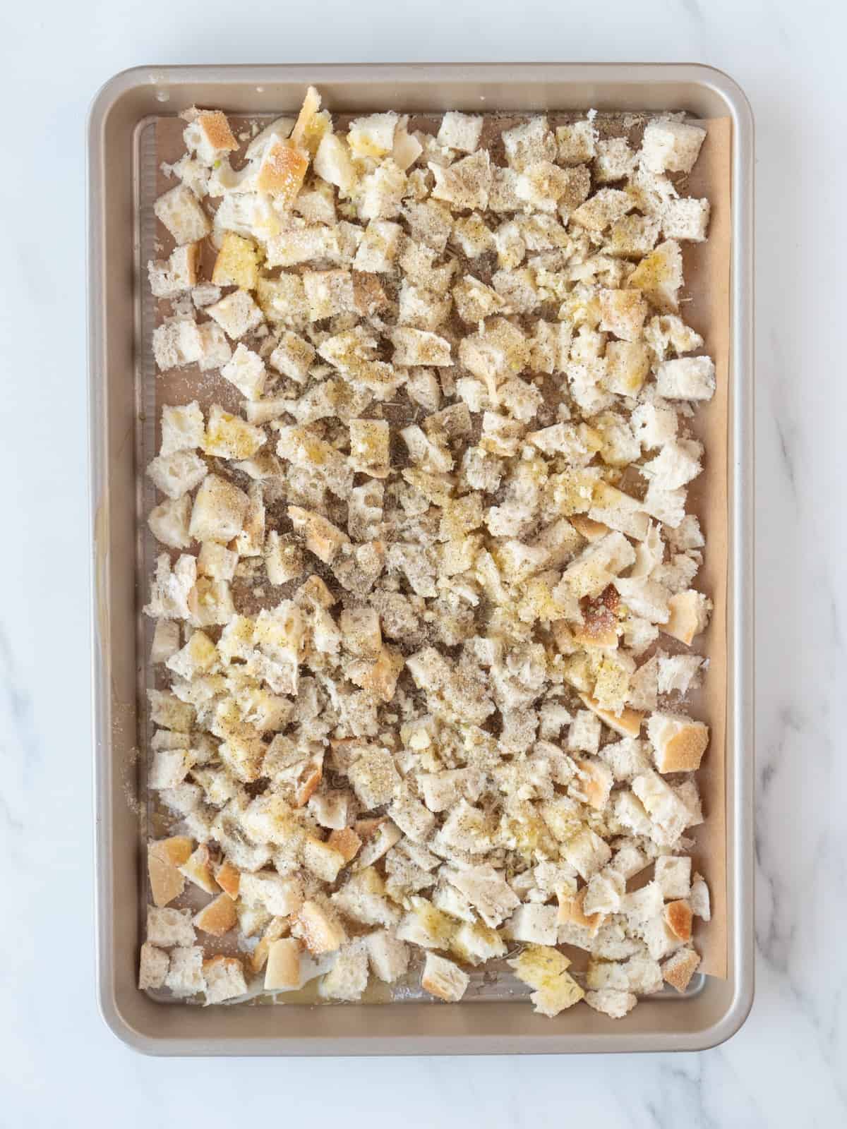 A rectangular parchment paper-lined baking sheet of cubed pieces of bread drizzled with olive oil and sprinkled with garlic, salt, pepper and italian seasoning.