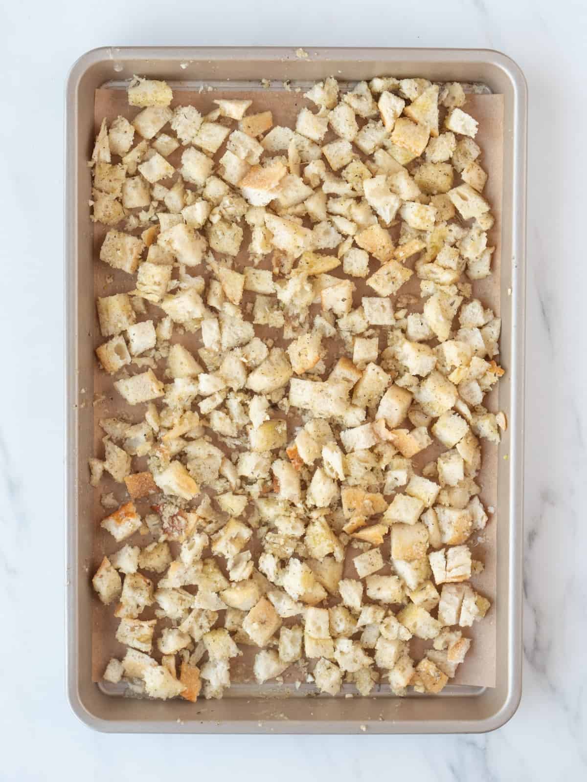 A rectangular parchment paper-lined baking sheet of cubed pieces of bread and tossed with olive oil and sprinkled with garlic, salt, pepper and italian seasoning.