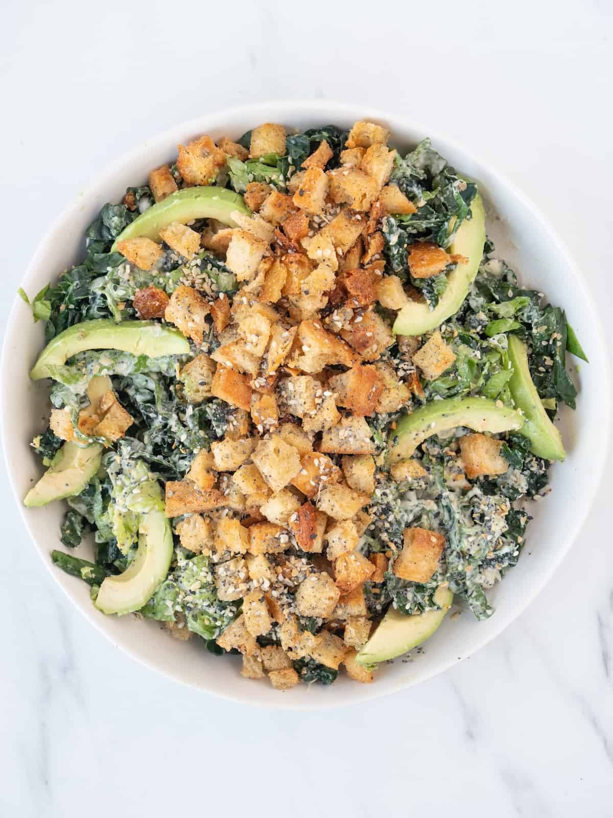 A low bowl with shredded lettuce and kale, tossed with caesar dressing, with avocado wedges and croutons on top, and a sprinkle of everything but the bagel seasoning.