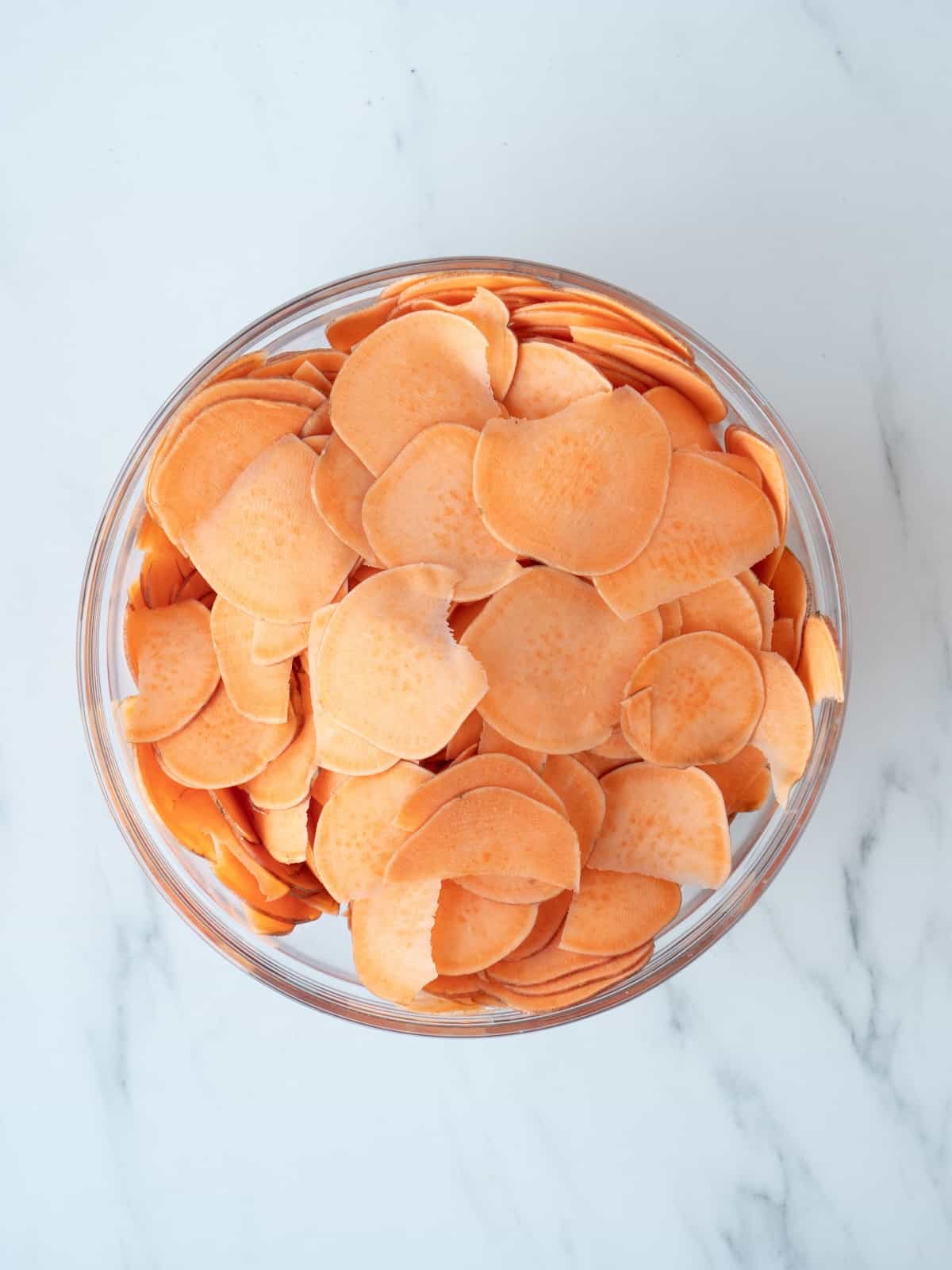 A large glass mixing bowl with thinly sliced sweet potato chips.
