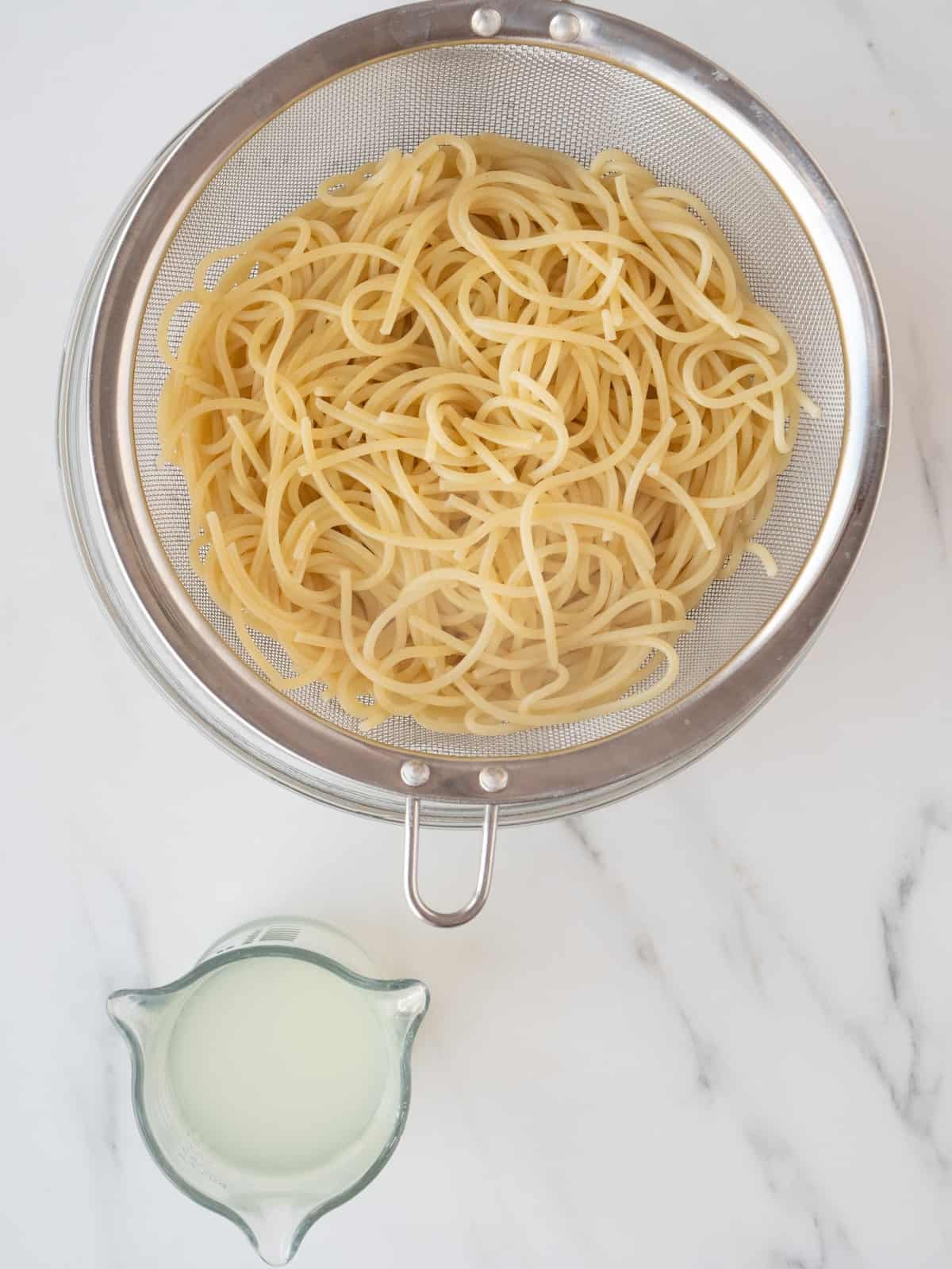 A glass mixing bowl with a large sieve with cooked spaghetti and a measuring glass with pasta water on the side.