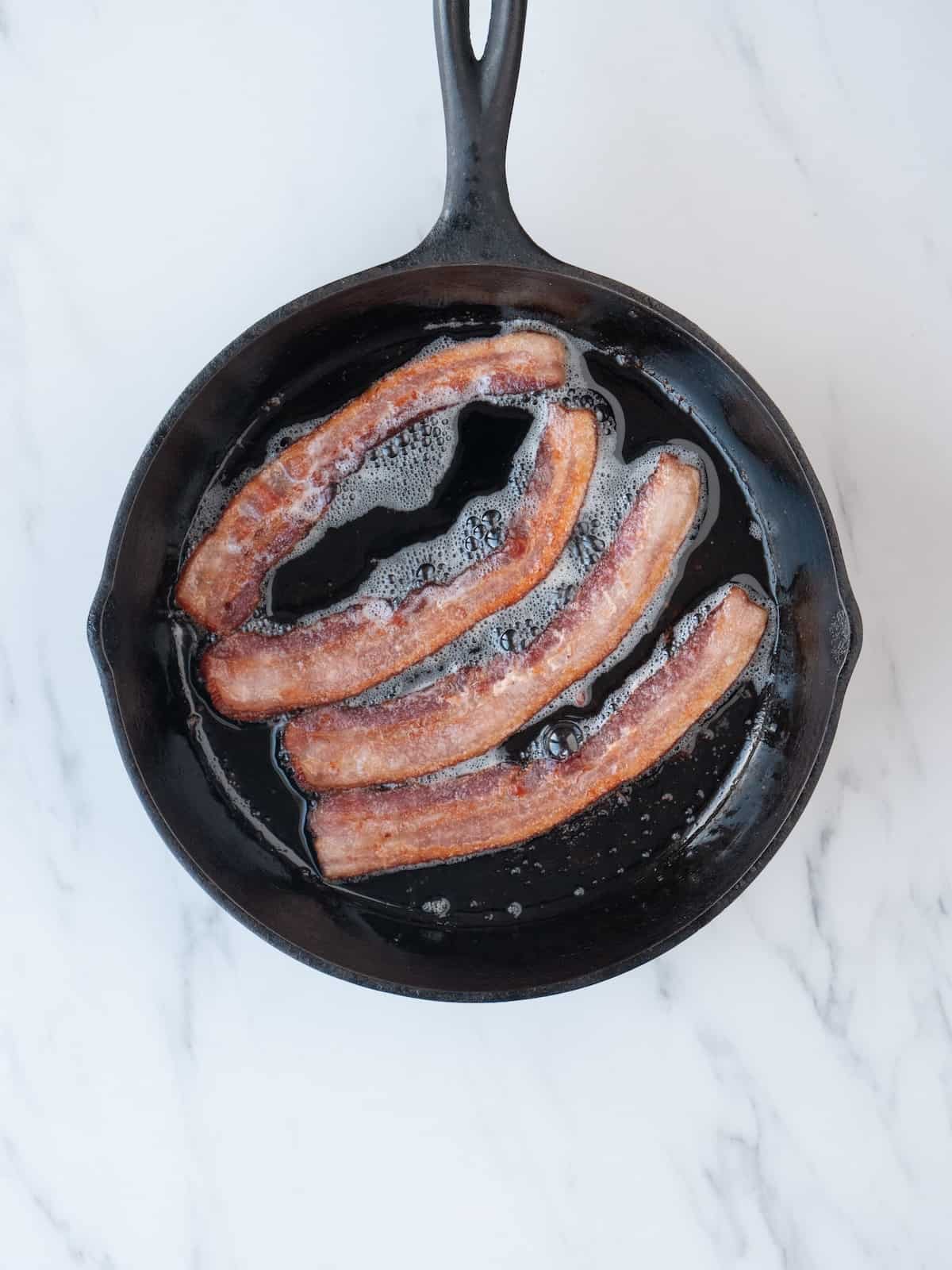 A skillet with thinly sliced bacon being cooked.