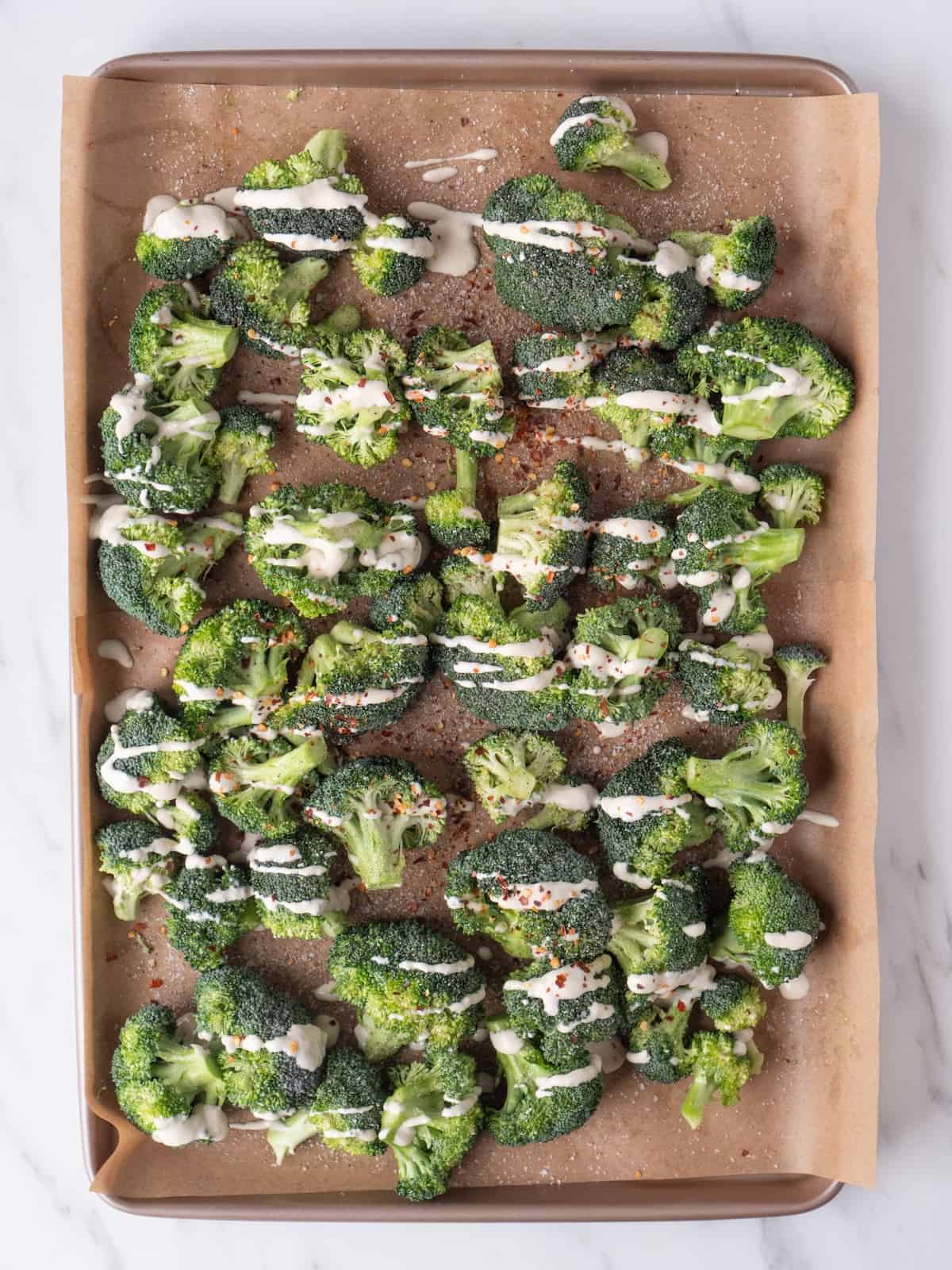A parchment-lined baking sheet with broccoli florets spread out evenly in a single layer tossed in oil, salt, pepper and red pepper flakes, with a drizzle of tahini sauce, made with lemon and garlic.