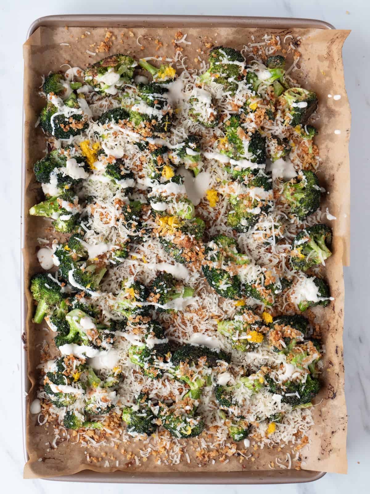 A parchment-lined baking sheet with broccoli florets baked, crispy and just out of the oven, topped with tahini sauce drizzle, toasted buttered breadcrumbs, lemon juice and zest and grated parmesan,.