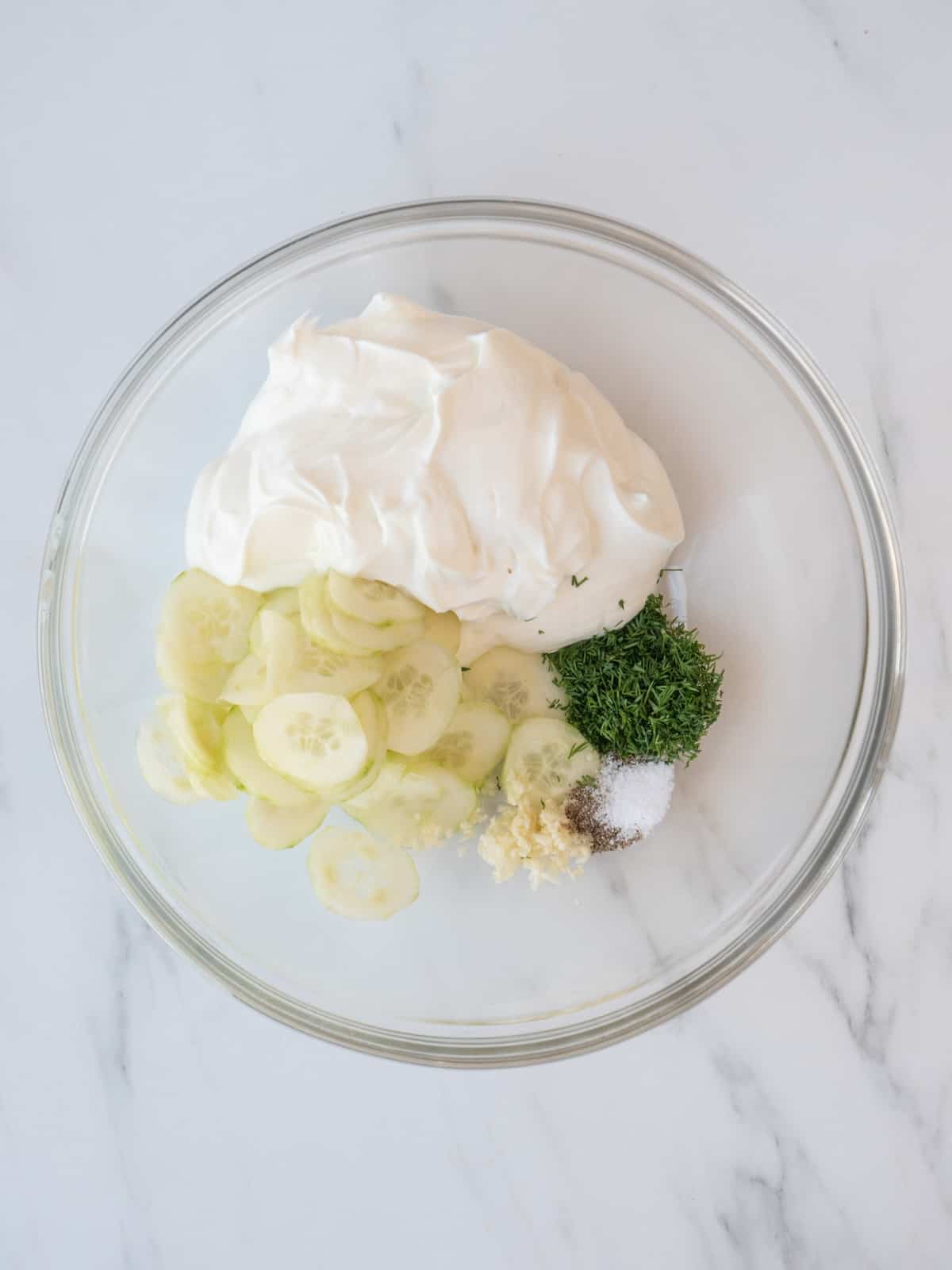 A glass mixing bowl with greek yogurt, sliced cucumbers, chopped dill, finely chopped garlic, and salt and pepper.