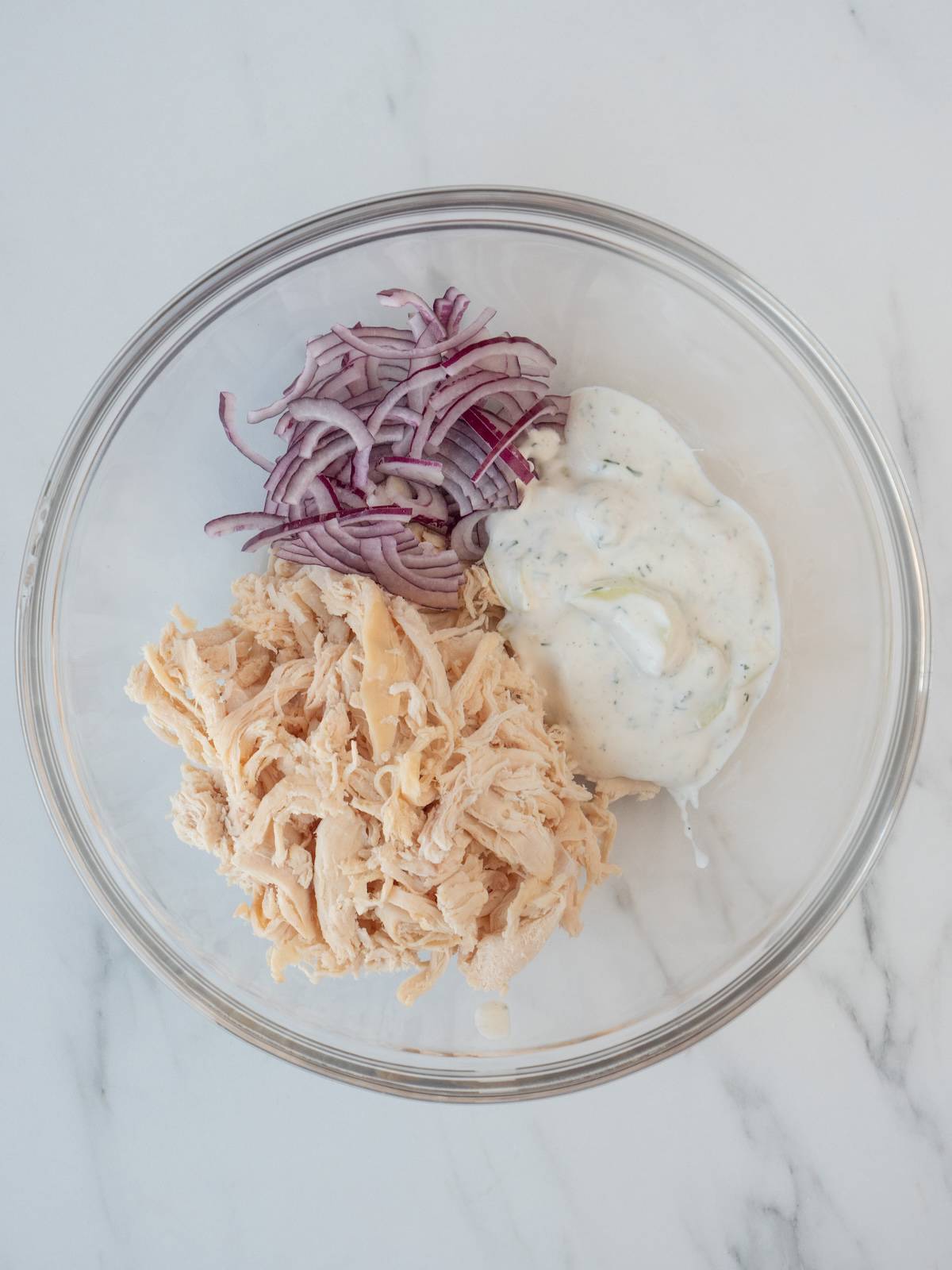 A glass mixing bowl with shredded chicken, thinly sliced red onions and tzatziki.