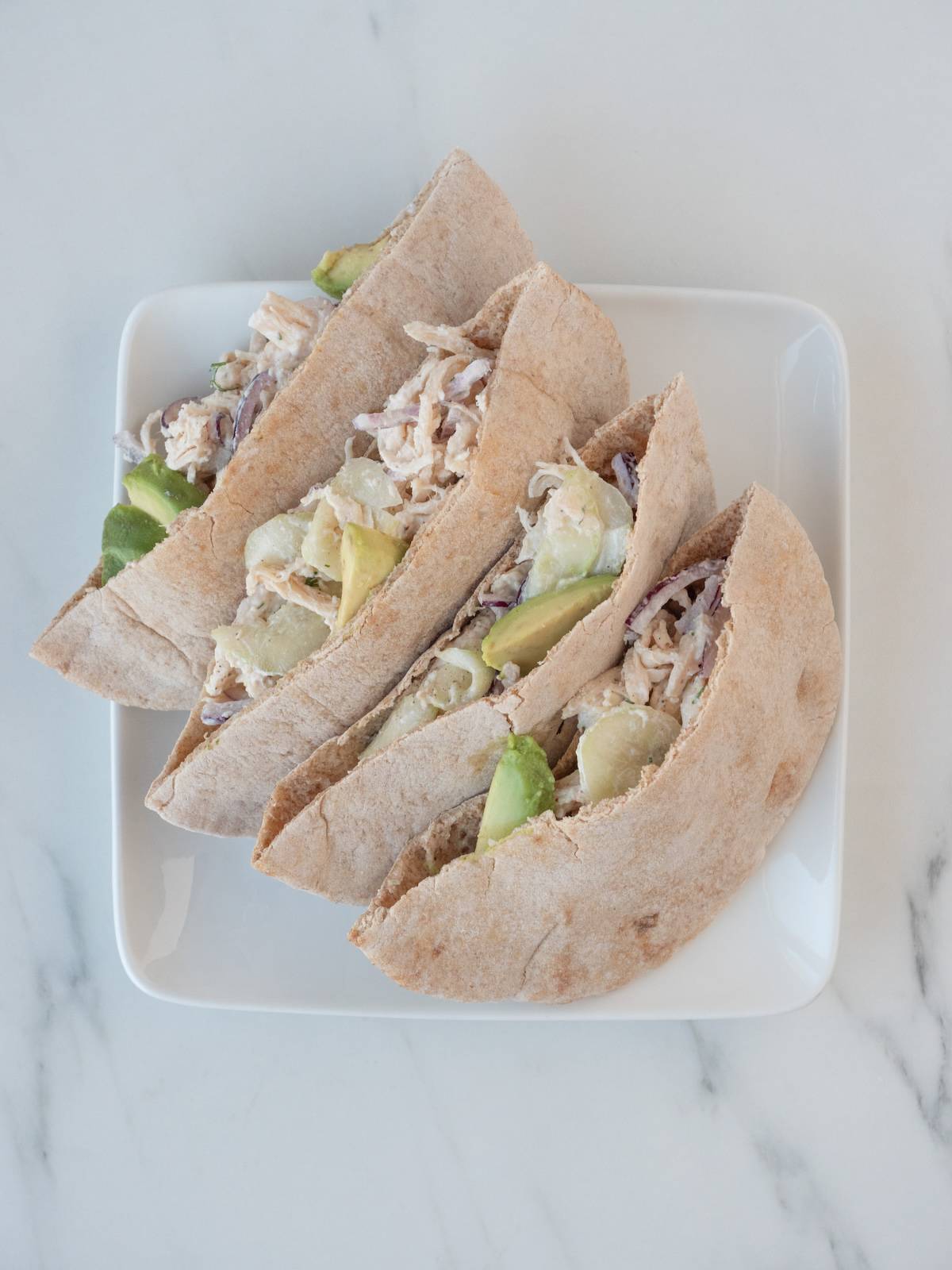 A square white plate, with four halves of stuffed pitas, stuffed with shredded chicken tossed in tzatziki, sliced red onions and sliced avocados.