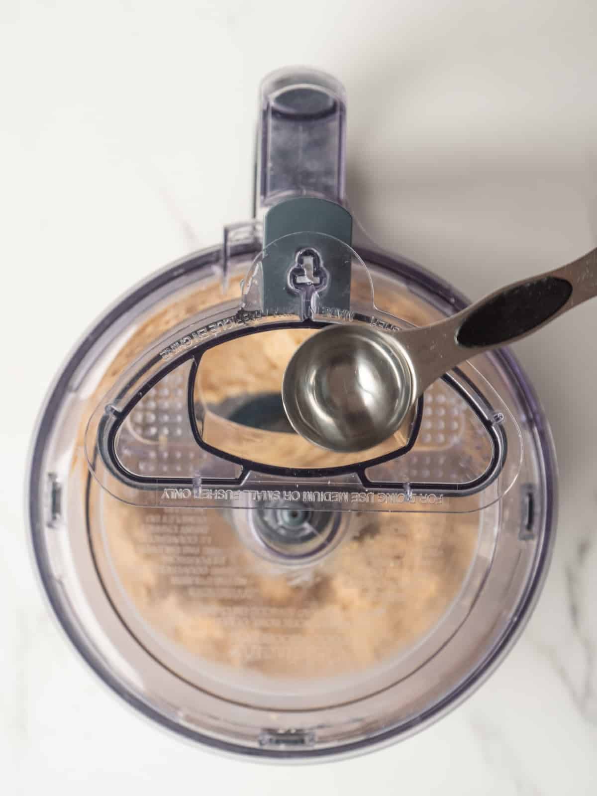 A food processor being run to make hummus, and a spoon being used to drizzle cold water from the top opening in the lid, with the food processor running.