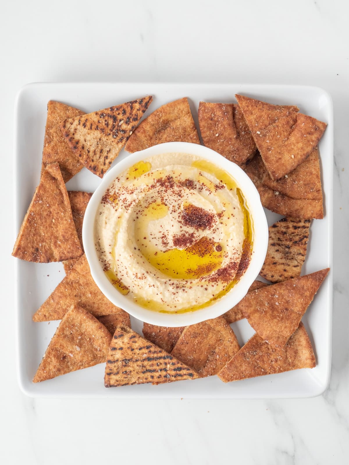 A square white platter with the baked pita chips dusted with za'atar and salt, and a bowl of hummus in the center that is garnished with sumac and olive oil.