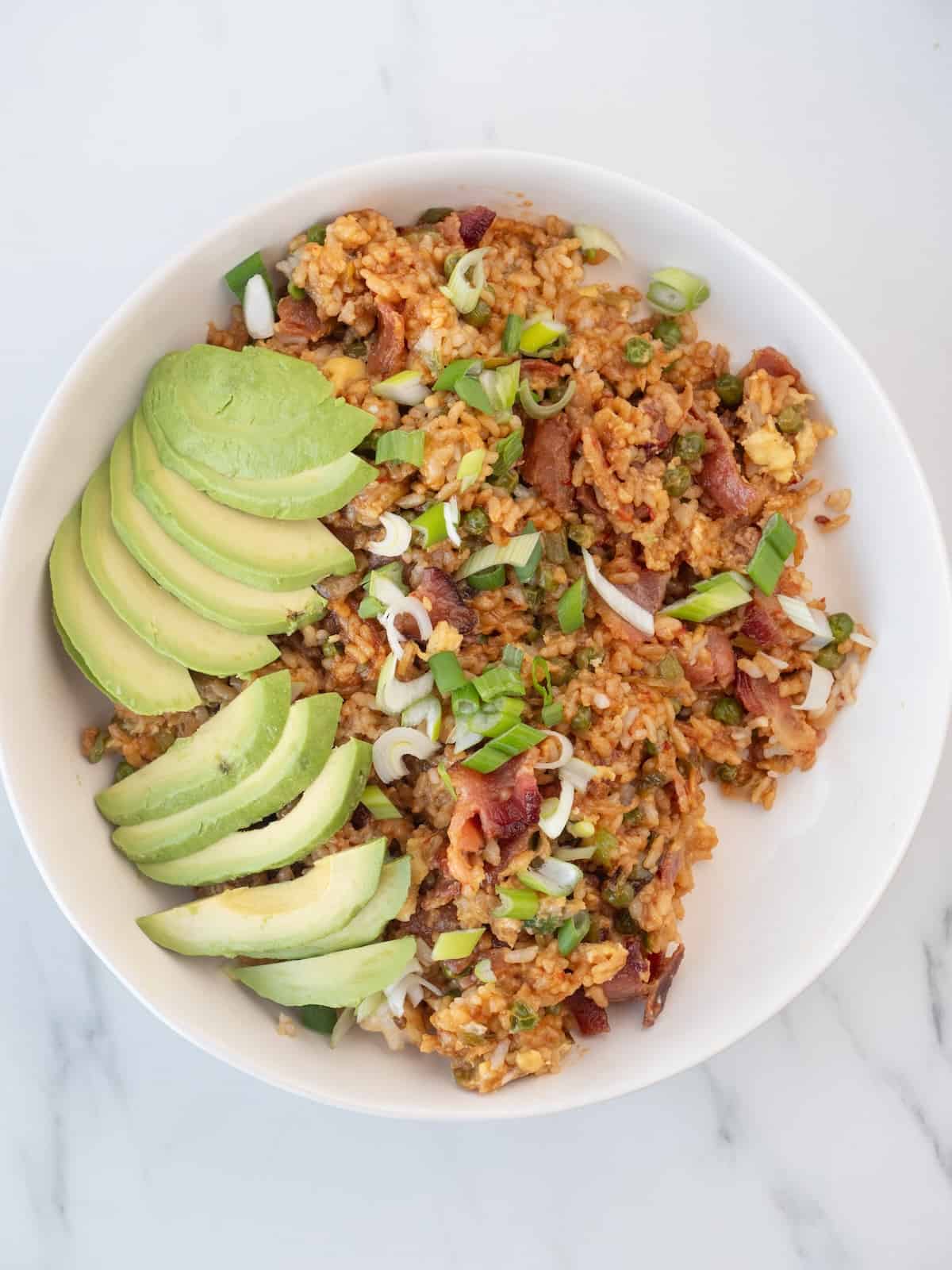 A bowl with spicy bacon fried rice, topped with avocado wedges and green onions.