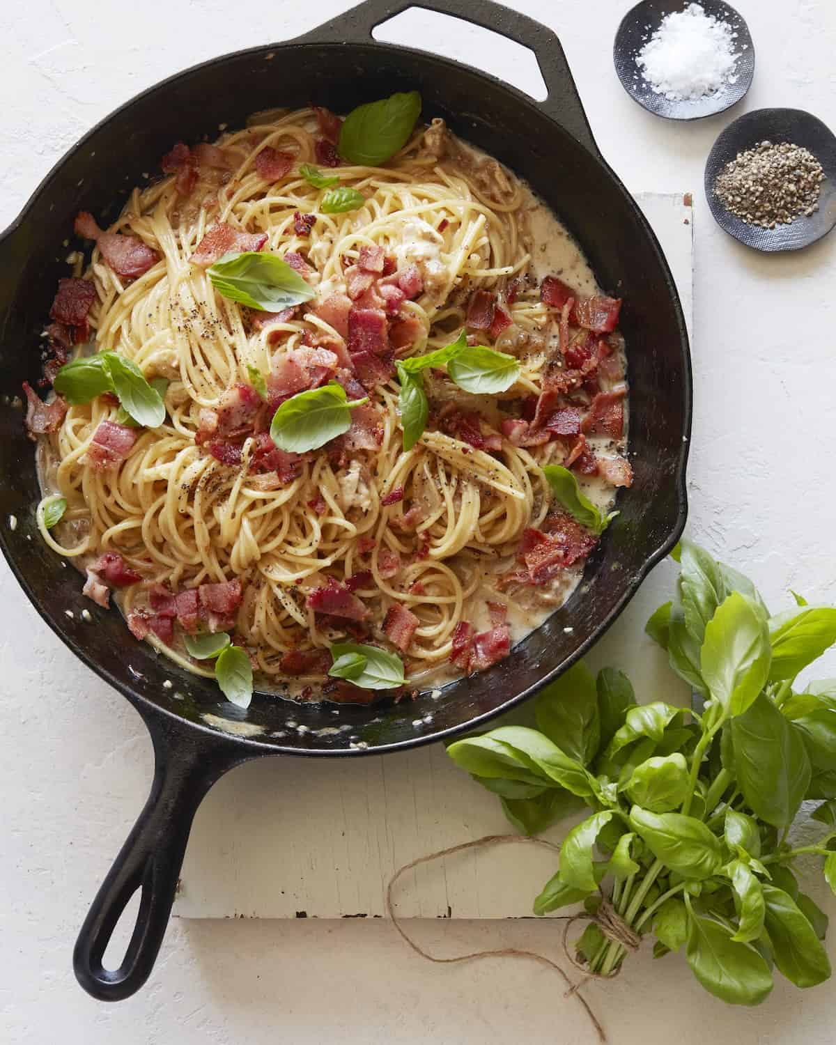 A skillet with brie, bacon and basil pasta with small bowls of salt and black pepper and basil.