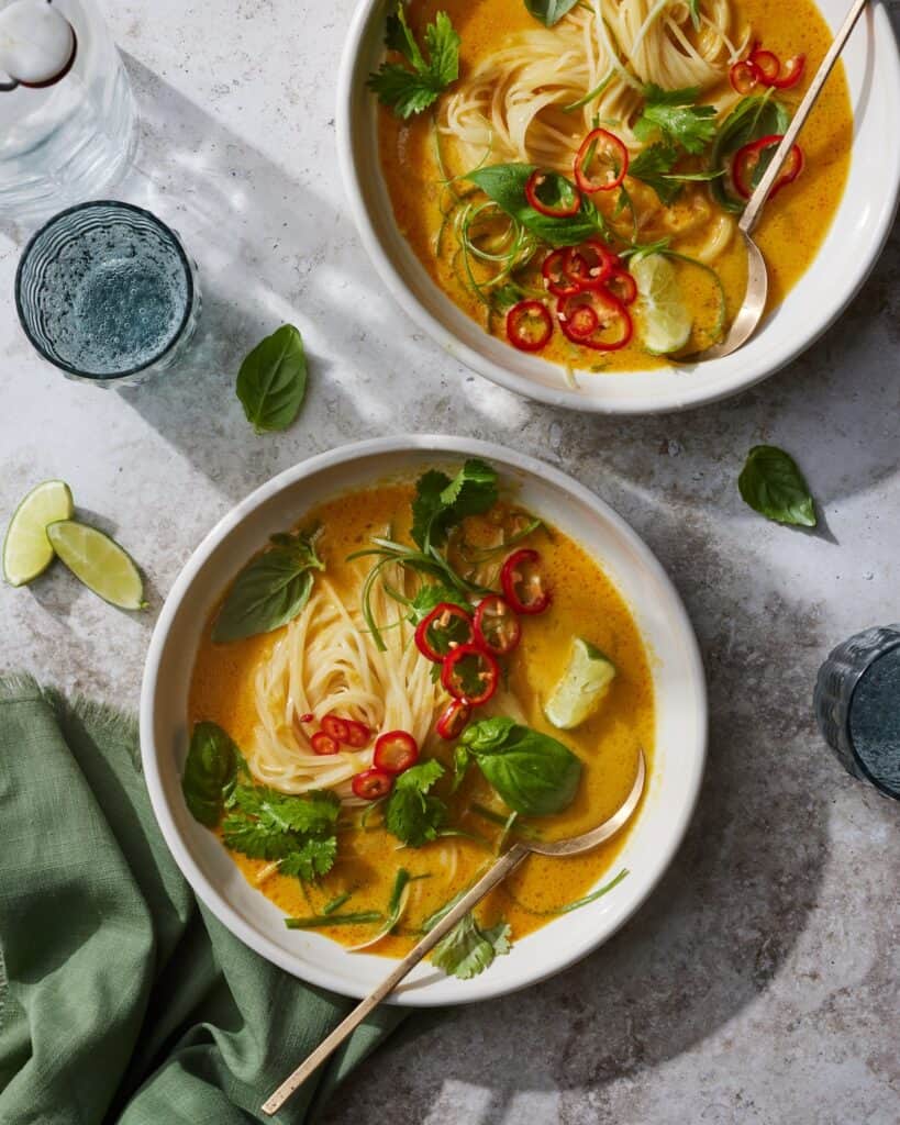 Two ceramic bowls with spoon with curry coconut noodle soup garnished with basil, fresno chili, green onion, cilantro and limes, with a drink, a green dish cloth and a few leaves of basil and lime wedges all placed around it.