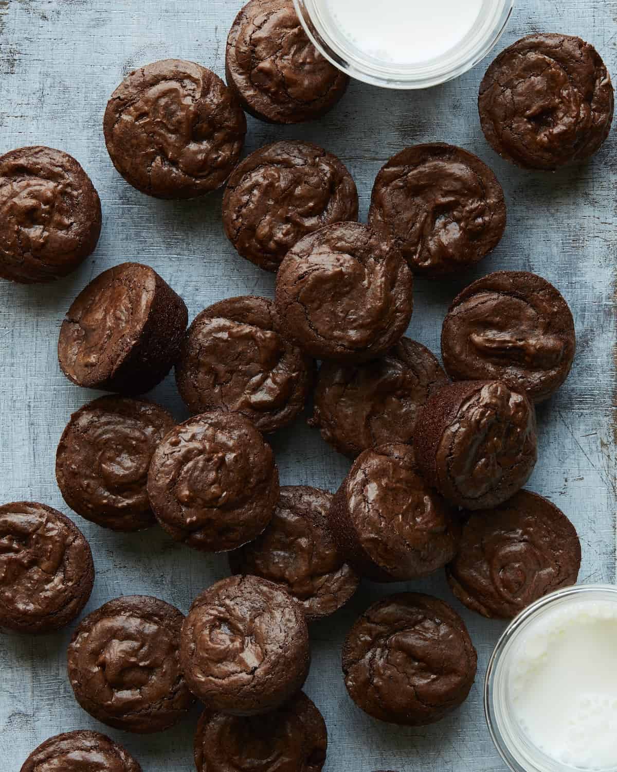 An overhead shot of crinkle top brownie bites placed on a blue counter with two small bowls of milk.