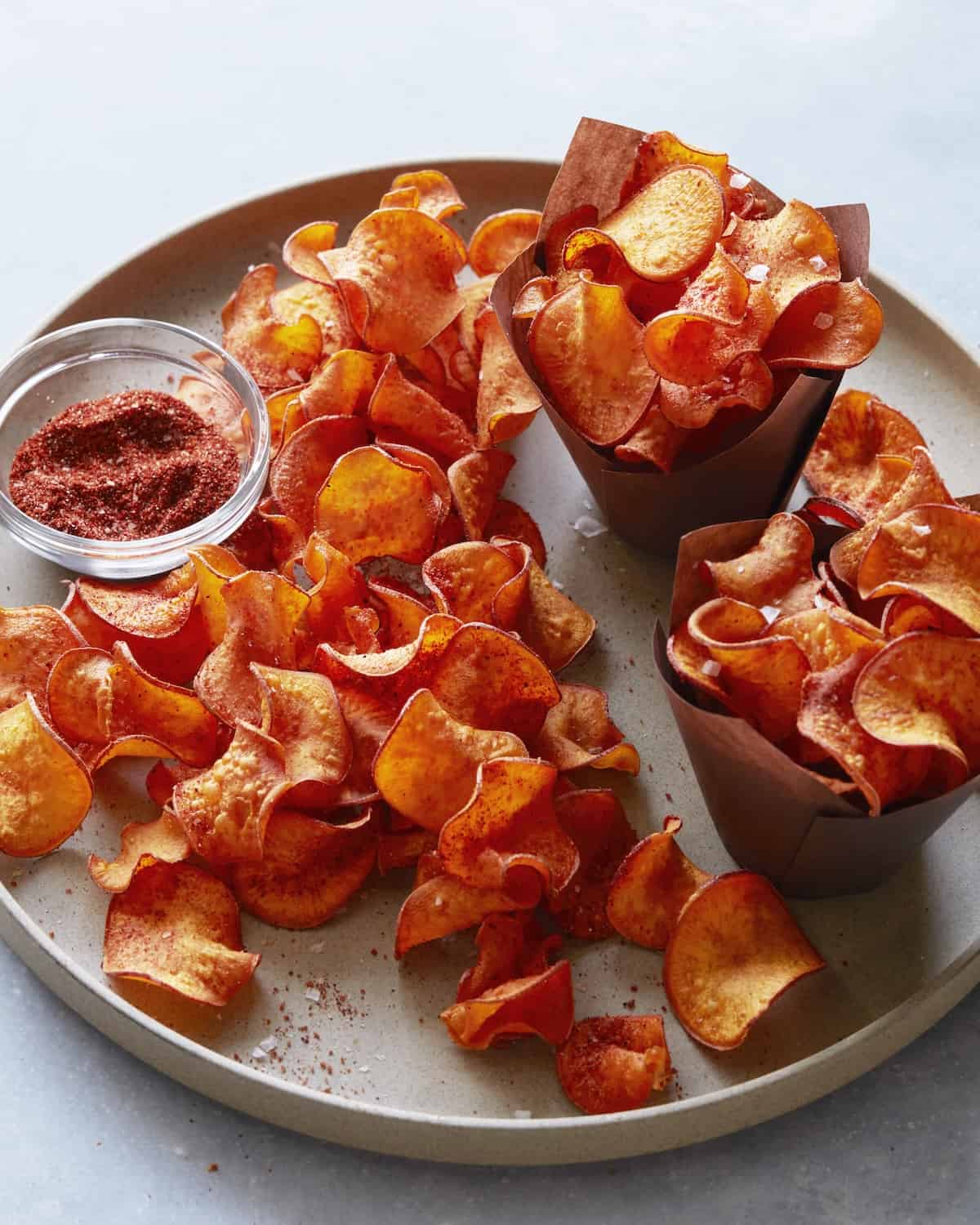 A round platter with BBQ sweet potato chips on the plate, along with two brown paper bowls filled with more of the chips, along with a small glass bowl of BBQ spice mix.