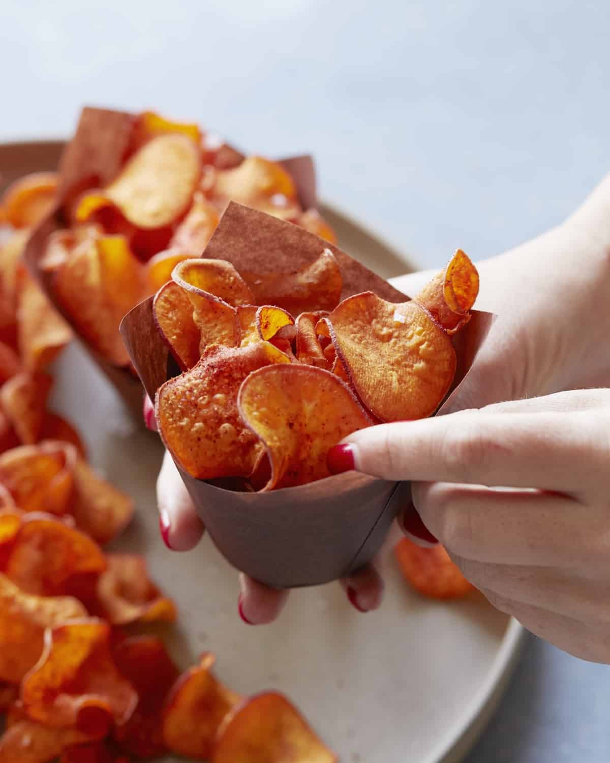 A woman's hand holding BBQ sweet potato chips filled in a brown paper bowl, with more chips in the background.
