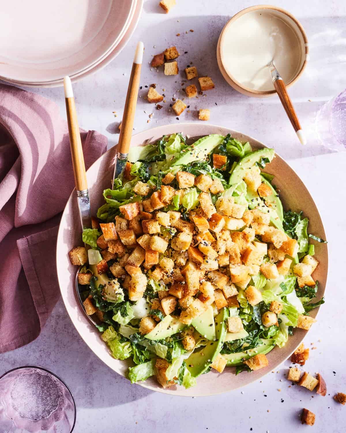 A low bowl with an avocado kale caesar salad, with some extra dressing in a small bowl on the side, and a pink kitchen towel next to it.