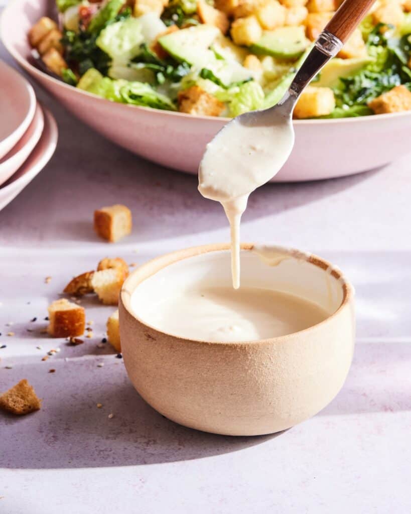 A closeup shot of a bowl of caesar dressing, with a spoon drizzling it into the bowl, with the blurry view of avocado kale caesar salad in the back.