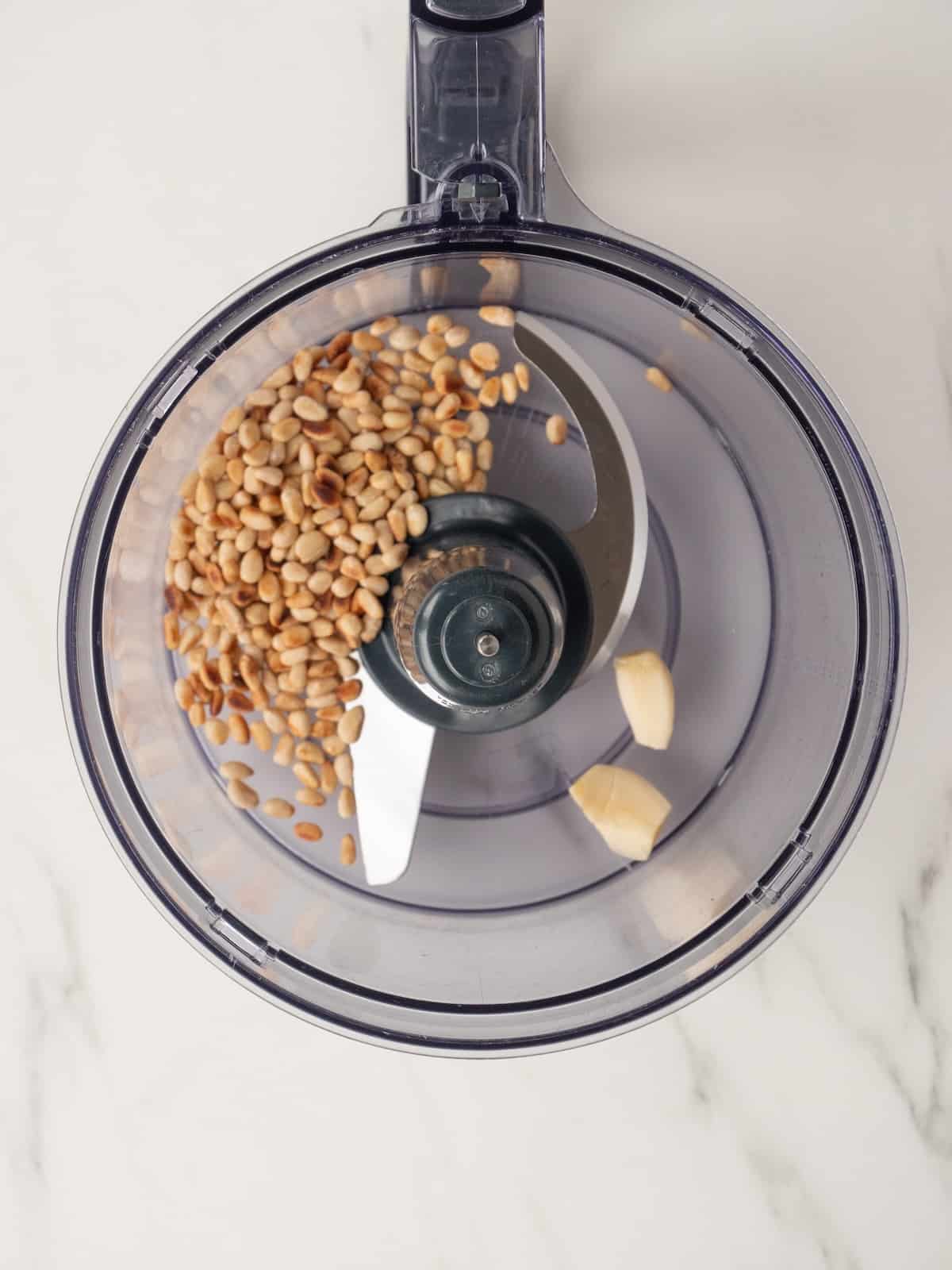 An overhead shot of a food processor with toasted pine nuts and garlic.