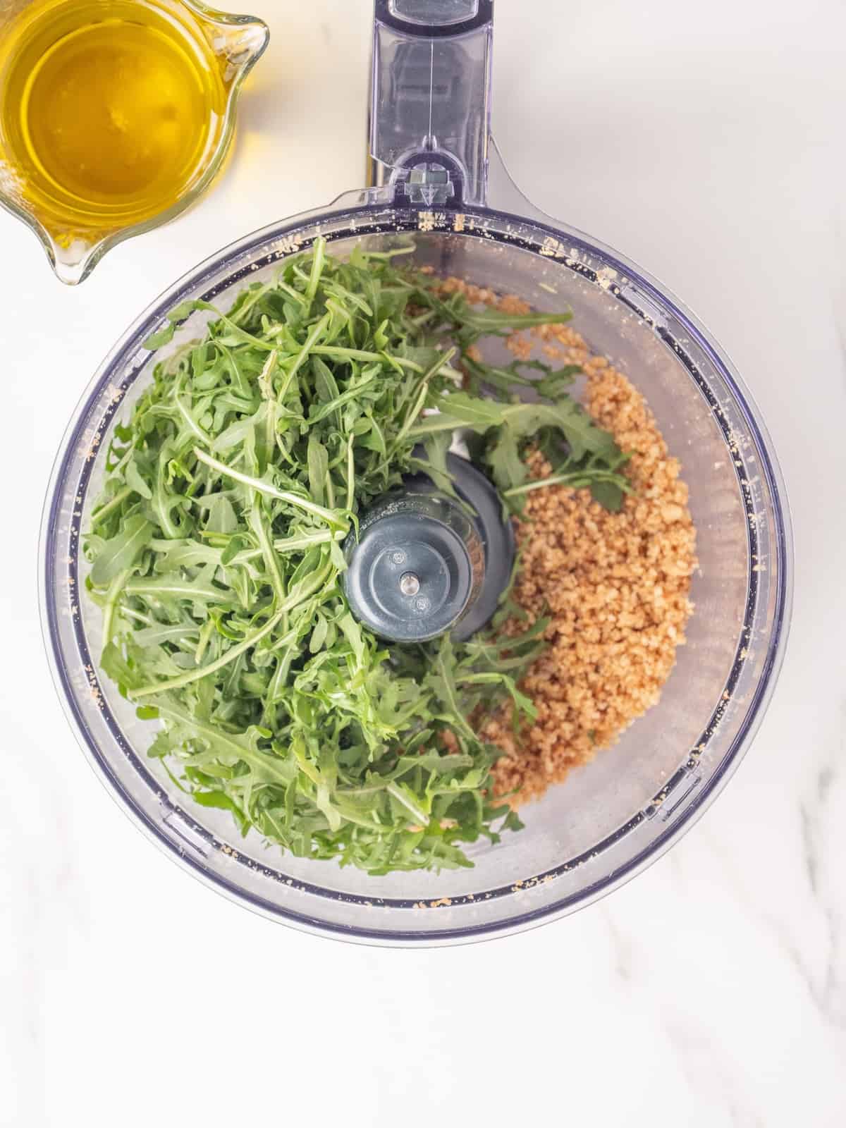 An overhead shot of a food processor with toasted pine nuts and garlic blended and fresh arugula added, along with a measuring cup with a spout containing olive oil.