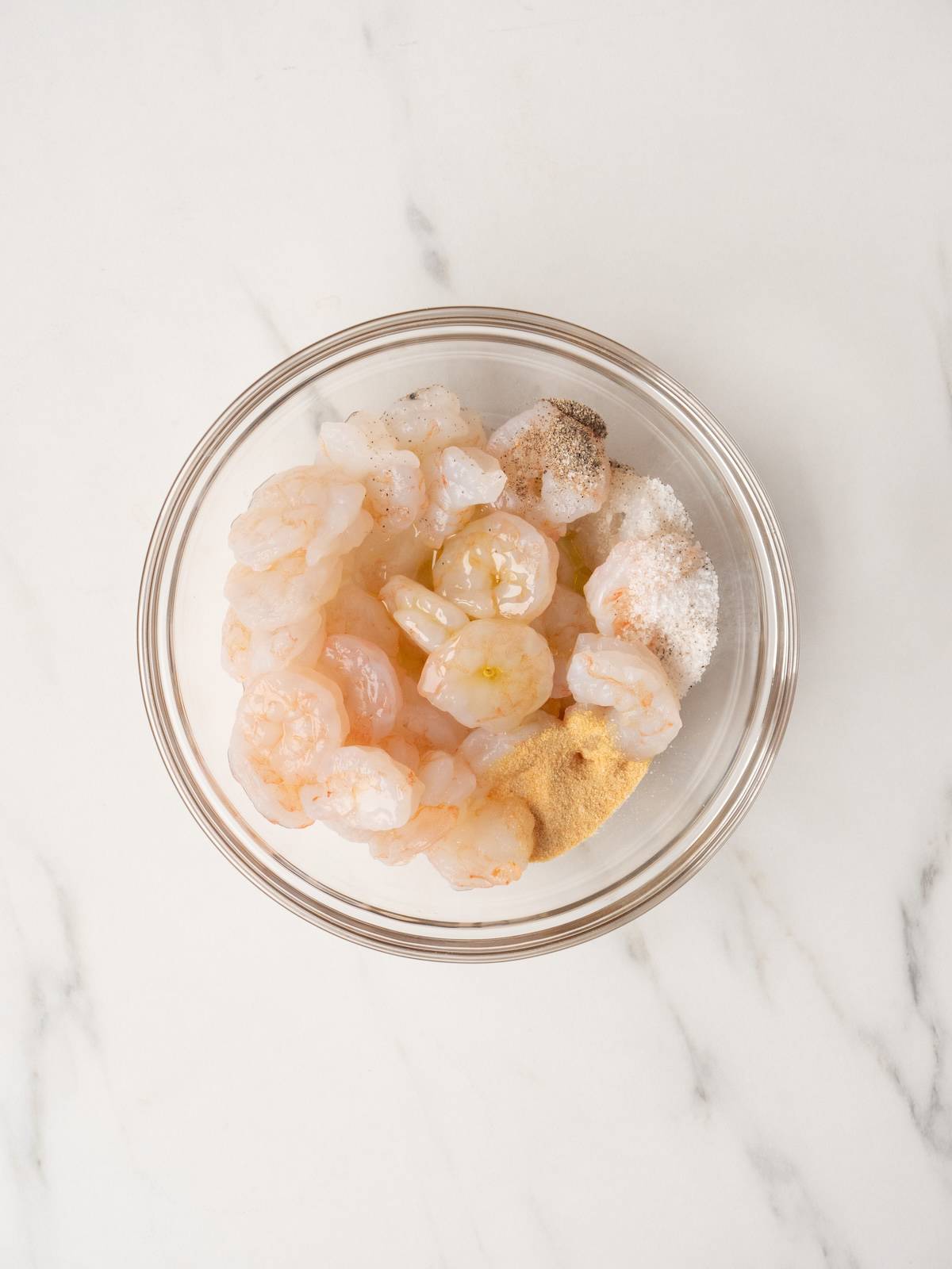 A small glass bowl with shrimp tossed with oil, garlic powder, salt and pepper.